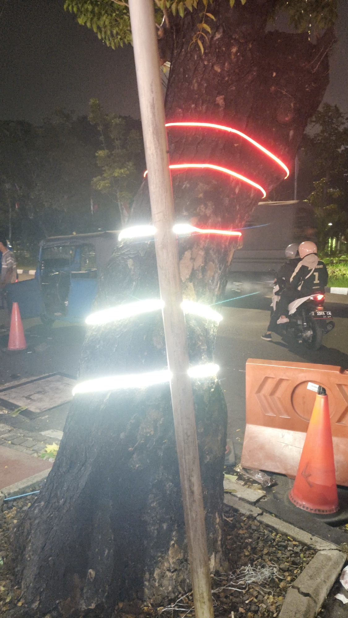 The image depicts a tree trunk adorned with multiple bands of red neon light and reflective tape, wrapped around it. The tree stands on a street at night, with a dark sky and trees visible in the background. In the foreground, a light-colored pole is positioned in front of the tree. To the left, a pink traffic cone and a blue vehicle are visible. Further to the right, two individuals on a motorcycle are riding on the street, and an orange traffic barrier with a cone is also present. The scene appears to be in a moderately lit urban environment. The license plate on the motorcycle reads "B 3385 CN". Based on the license plate format, the location is likely in Indonesia, specifically Central Jakarta, Indonesia.