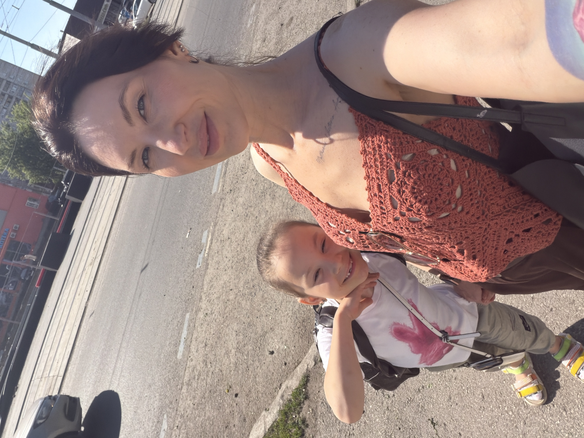 The image presents a selfie taken outdoors in Perm, Russia. The focal point is a woman and a child. The woman has dark hair and fair skin, wearing a dark strap over her shoulder and an orange crocheted top. A faded tattoo is visible on her upper chest. The child, slightly in the foreground, has light skin and is wearing a white t-shirt with a faded graphic and khaki pants. The child also has visible white sandals with yellow and green accents.
The background depicts a cityscape. Specifically, a street with tram tracks and a red and white building, identified by signs in Russian script. The lighting indicates it is daytime.