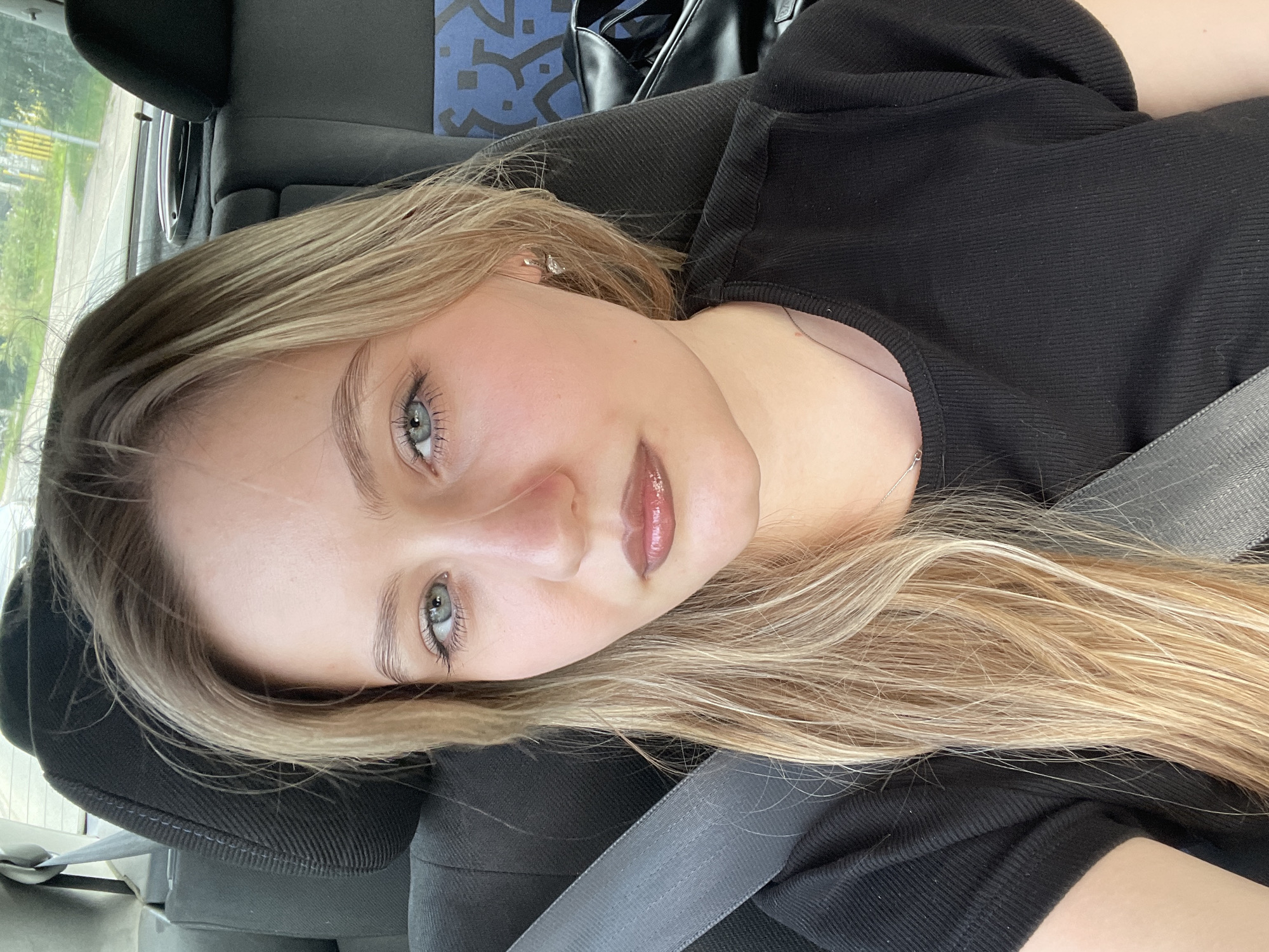A young woman with blonde hair and blue eyes is seen in the passenger seat of a car, wearing a black t-shirt and a seatbelt. The interior of the car is visible, including the headrest and part of the back seat. The woman is looking directly at the camera, with her makeup including eyeshadow, mascara, and lipstick. A blurred background shows a street and greenery, suggesting the car is in motion within an urban or suburban setting.