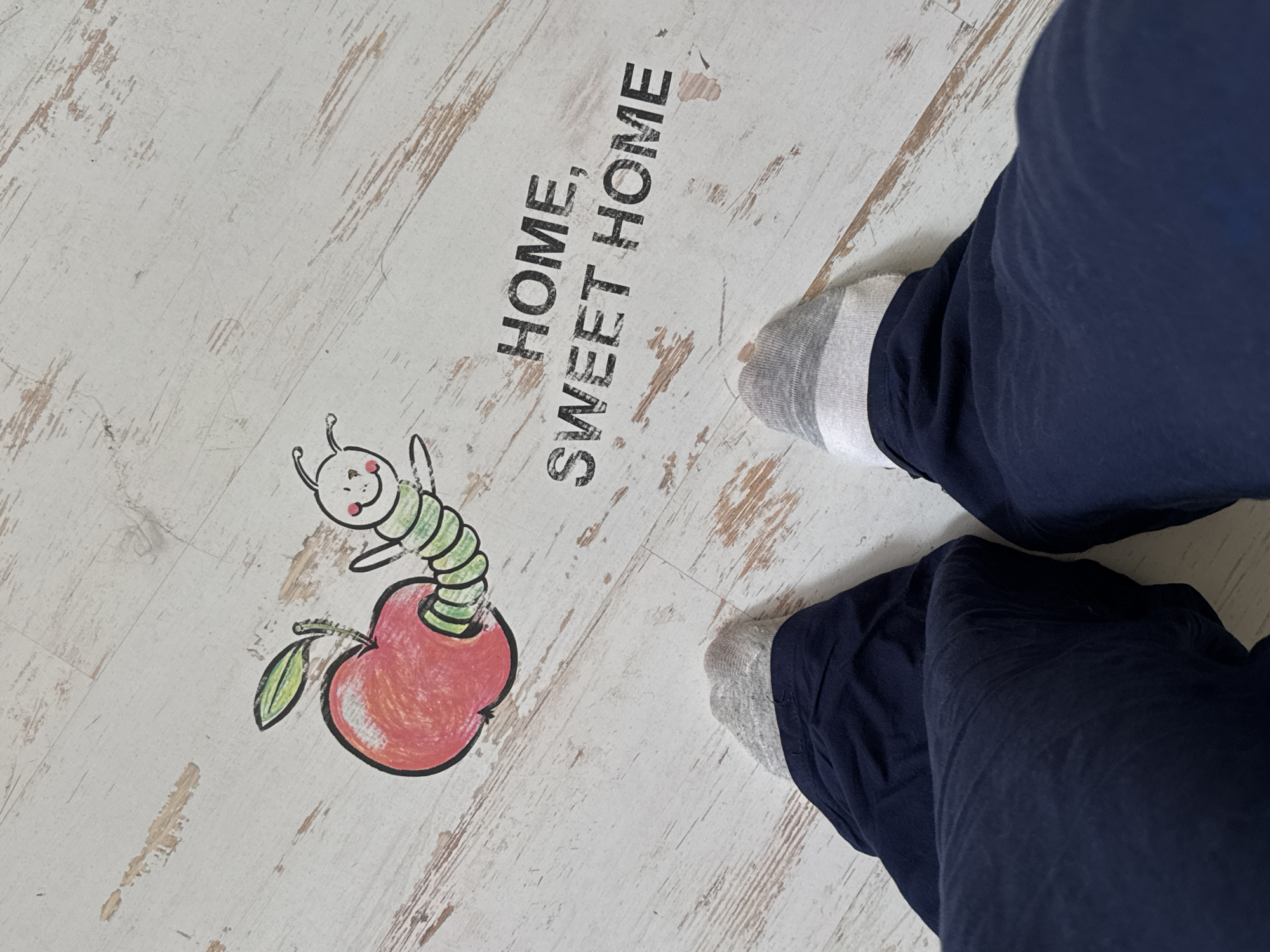 The image shows a close-up view of a person's legs and feet wearing navy blue pants and grey socks. They are standing on a white wooden floor with a distressed finish. A drawing of a caterpillar emerging from an apple is to the upper left of the frame. To the right, the words "HOME, SWEET HOME" are printed in black capital letters. The scene is indoors, likely in a residential setting in Kolomna, Russia, given the context. The perspective is from a first-person point of view, looking down at the subject's own feet.