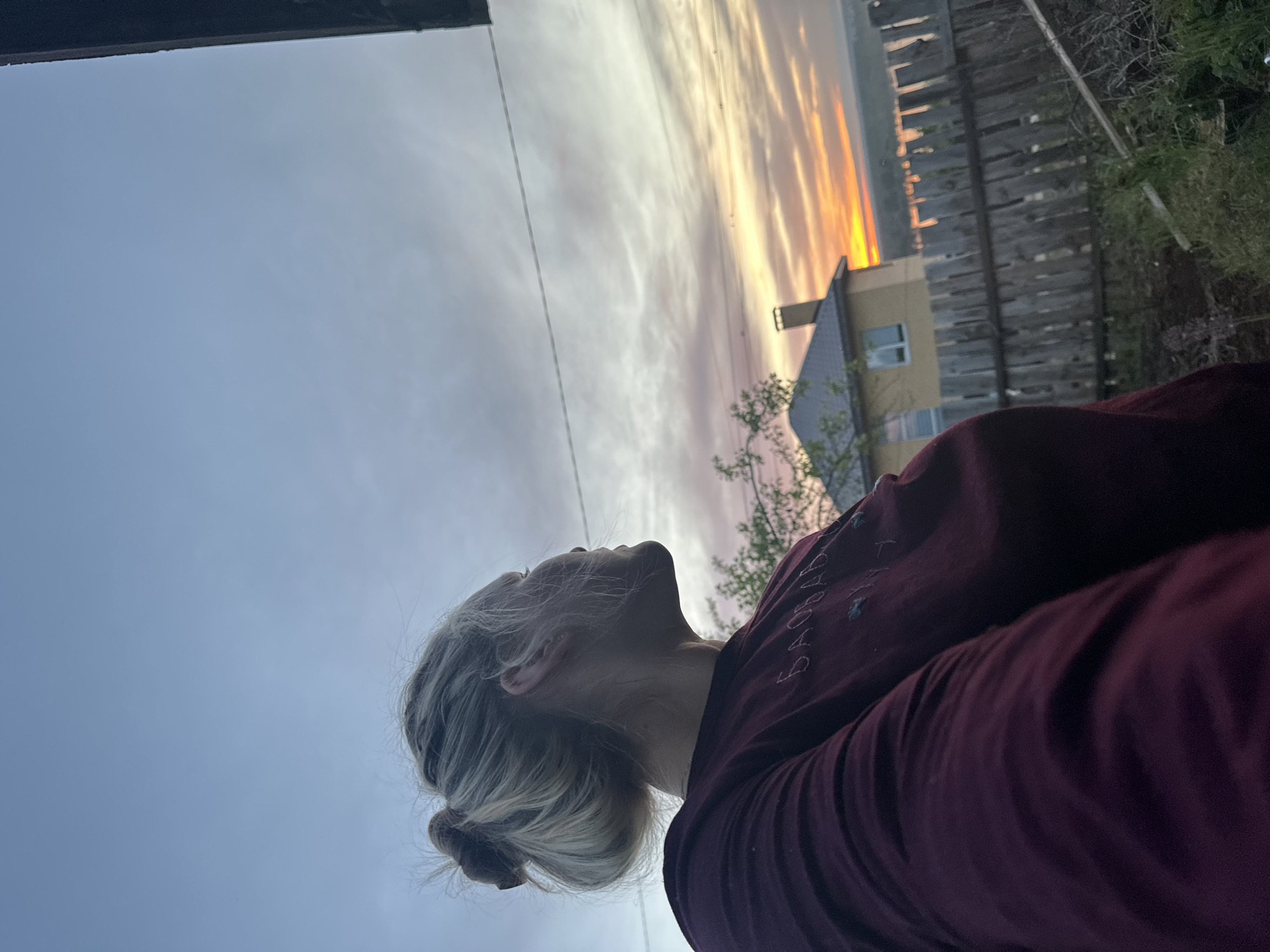 The image shows a woman with blonde hair pulled back into a bun, looking upwards. She is wearing a dark maroon sweatshirt. In the background, there is a fence, foliage, and a building with a visible window. The sky is visible, featuring a sunset with orange and grey clouds. The location is Chaikovsky, Russia.