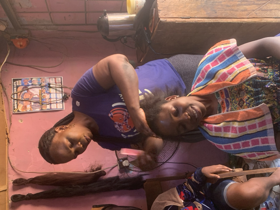 The image captures an indoor scene in Garaku, Nigeria, depicting a hair styling activity within what appears to be a small salon or a home-based styling area. A dark-skinned adult female, the stylist, stands wearing a purple short-sleeved t-shirt and grey bottoms, her hair in braids. She is actively working on the hair of a seated dark-skinned adult female client. The stylist holds a pink comb in her left hand, with both hands manipulating the client's natural, unbraided hair. The client wears a multi-colored striped towel draped over her shoulders and a patterned top, looking downwards with a slight smile. A third individual is partially visible on the far left, seated on a wooden chair, wearing a patterned top and shorts.
The room features light pink walls. To the left of the stylist, two bundles of dark hair extensions hang. A black oscillating fan is positioned behind the stylist. On a wooden surface to the right, a silver electric kettle, a yellow lidded jar, and black hair styling tools including a flat iron and combs are arranged. A calendar-style poster titled "HAPPY MARRIED LIFE." with multiple images is affixed to the wall, above which exposed electrical wiring is visible. Natural light enters the space, casting highlights and shadows.