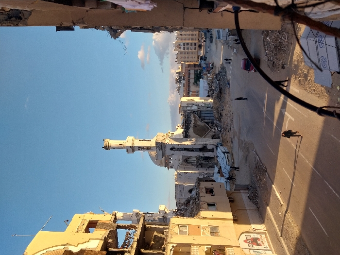 This image captures a heavily damaged urban street scene in Khan Yunis, Palestinian Territory, under a clear blue sky with sparse clouds. The central element is a prominent mosque, white in color, featuring a dome and a tall minaret, which exhibits extensive structural damage but remains partially upright.
To the left, a multi-story building, likely residential or commercial, is severely compromised, with large sections of its facade collapsed, exposing interior rooms and structural rebar. A prominent red and white sign with "SQUID GAME" graphics is affixed to its lower exterior. On the right, another multi-story structure displays significant destruction, revealing internal components and rubble, with articles of clothing visibly hanging from an upper section.
The asphalt street running through the middle foreground is partially clear, marked by some road lines. Both sides of the street are covered in extensive debris, including piles of concrete, twisted metal, and other building materials from collapsed structures. Several individuals are present: one person in dark clothing walks across the foreground street, casting a long shadow. Other smaller figures are observed further down the street, some near the mosque and others amidst the rubble. A red vehicle is visible on the right side of the street in the mid-ground. The scene is illuminated by the low sun, casting long shadows, suggesting either early morning or late afternoon.