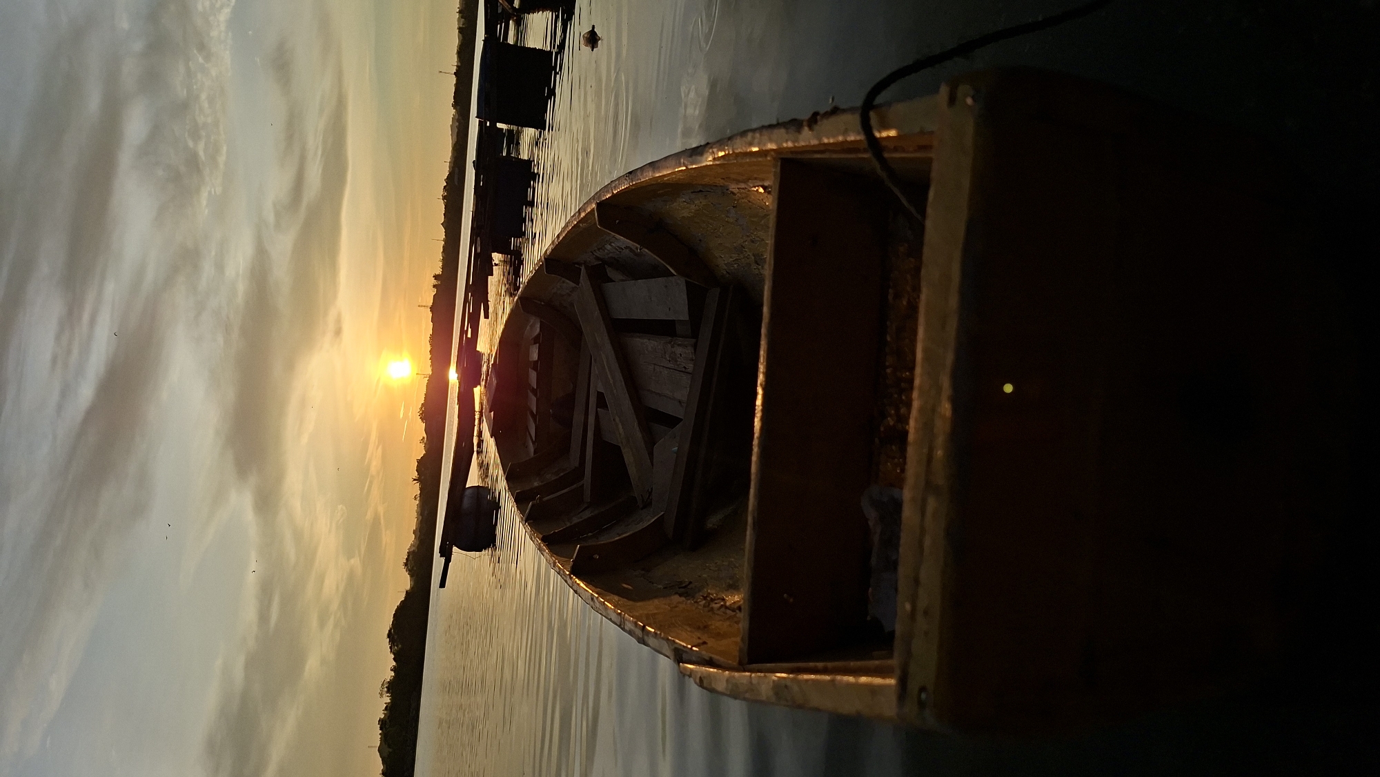 An outdoor vertical shot captures a wooden rowboat on calm water during sunset. The sun, a bright orb, is low on the horizon, casting a warm orange glow across the sky and reflecting on the water. Wispy clouds streak across the sky, tinged with the colors of sunset. The boat, seen from a low angle, is empty and contains loose wooden planks. In the background, a silhouette of trees and structures lines the distant shore, and several floating structures or buoys are visible in the water. The scene appears to be a serene waterfront setting, possibly a lake or a quiet bay. Based on the visual cues, this scene is likely located in Lhokseumawe, Indonesia, a coastal city known for its natural beauty and waterfront areas.