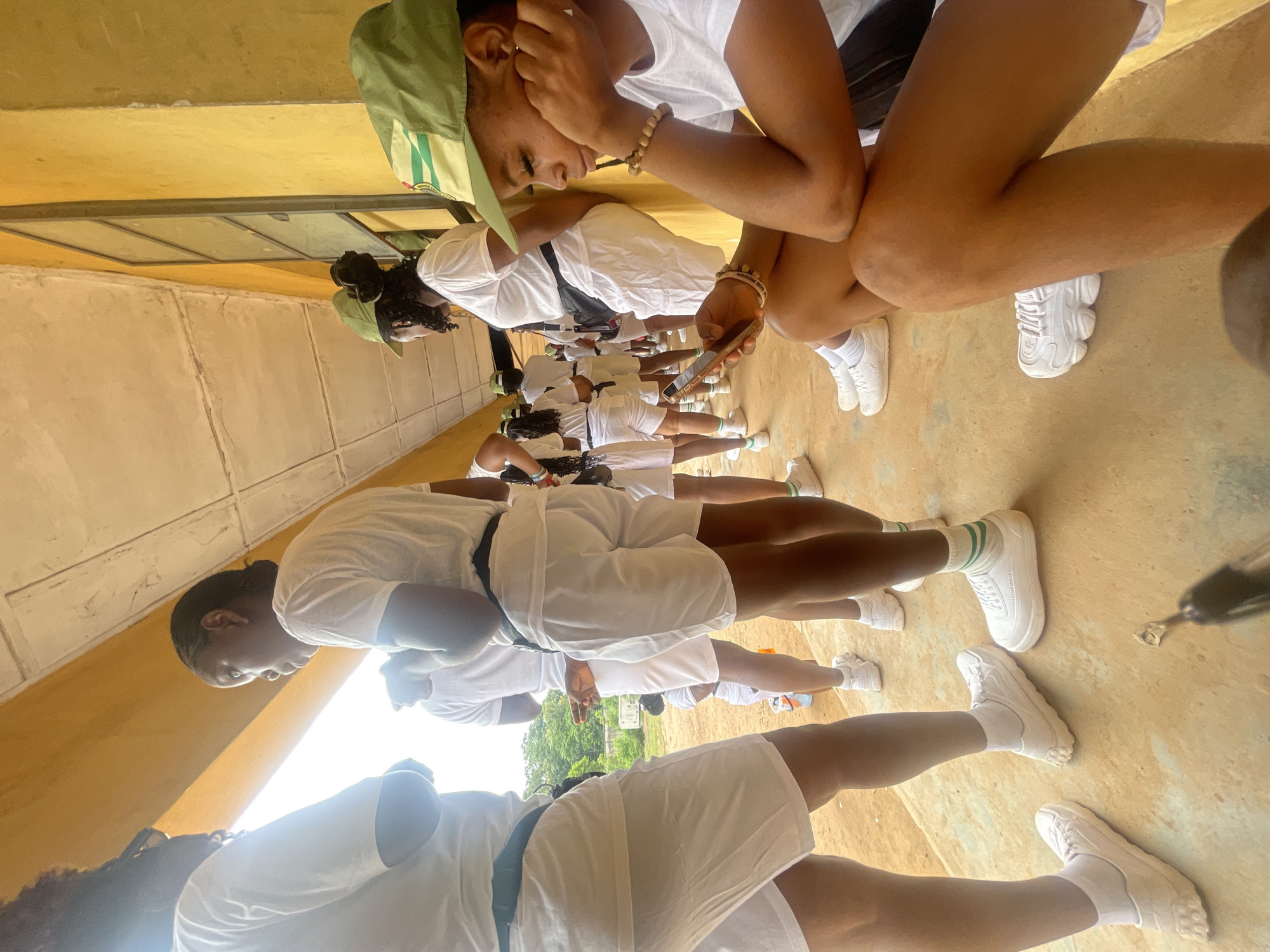 In Ise-Ekiti, Nigeria, a group of young people, likely participants in a youth corps program, are gathered outdoors under a yellow-painted building with a concrete overhang. The ground is a sandy, unpaved surface. Most individuals are wearing white t-shirts and white shorts, with some donning green caps.
In the foreground, a young woman is seated and looking at her mobile phone. She has dark skin, braided hair, and is wearing a white t-shirt, black shorts, white socks, and white sneakers. She has a beaded bracelet on her wrist. To her left, other individuals are standing, also in white attire. Some are looking forward, others are engaged in conversation. The scene appears to be during daylight hours, with natural light illuminating the area. The building's architecture suggests a functional, possibly institutional setting. The overall atmosphere is one of casual gathering and waiting.