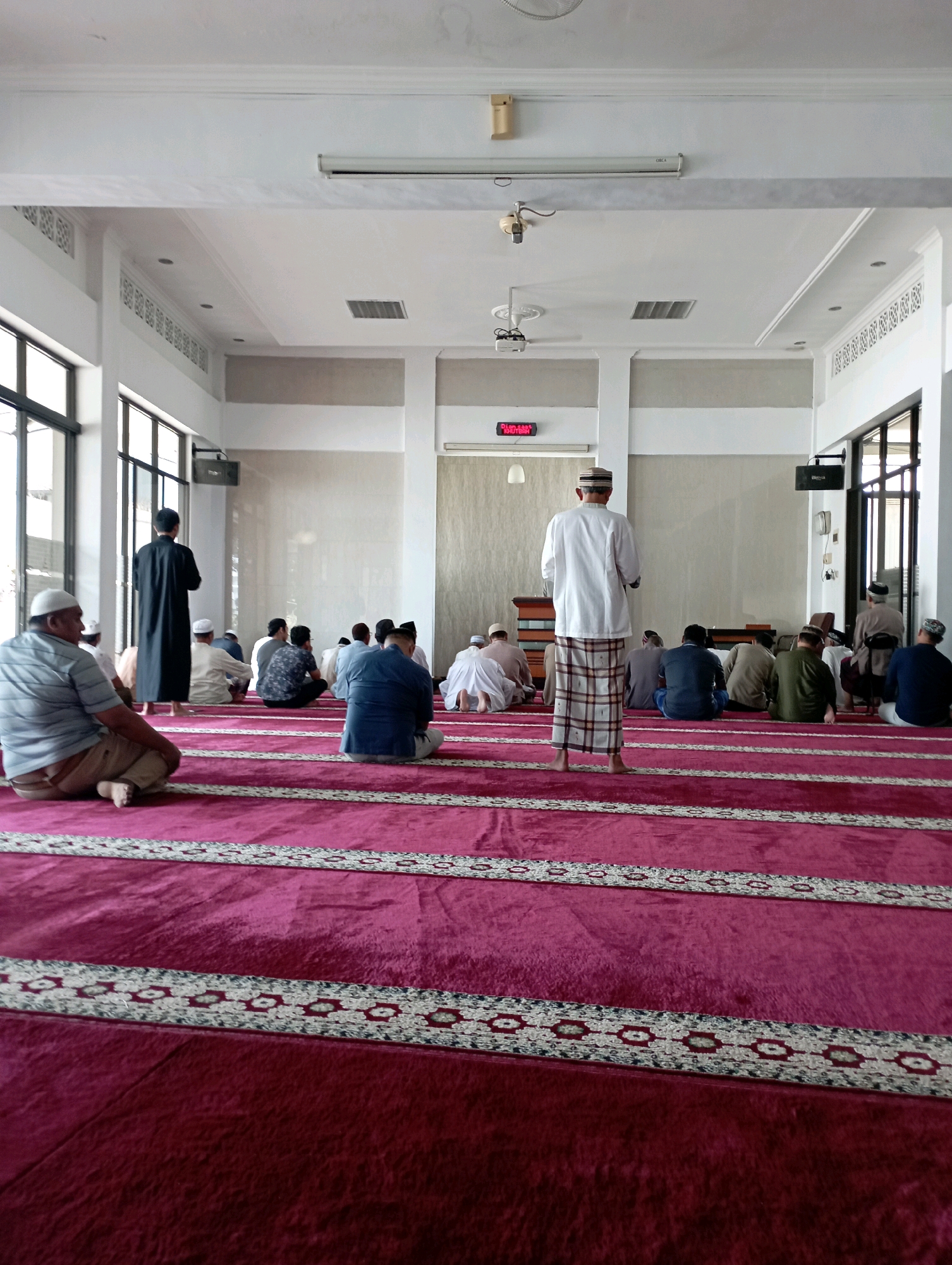 This is a vertical, eye-level, full shot of a mosque interior in Malang, Indonesia, during prayer. The floor is covered in a plush, deep pink carpet with white and silver patterned stripes marking prayer rows. In the foreground, a man in a grey and blue striped shirt and light brown pants sits on the carpet, facing away from the camera. To his left, another man in a black robe stands, facing the front of the mosque. The midground shows about a dozen men seated on the carpet, facing the front in prayer. A few men stand in the background, and others are visible in the far distance. The room has a high, white ceiling with recessed lighting and ventilation grates. Large windows with dark frames are visible on the left side of the frame, letting in natural light. The far wall is a muted cream color. A digital clock on the far wall displays red digits.