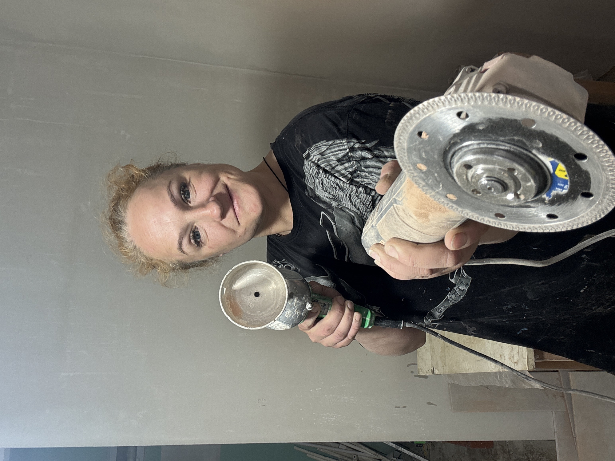 A woman holds two different angle grinders up to the camera. The location appears to be indoors in the city of Chaikovsky, Russia, near a wall that is partially painted. The woman is smiling slightly at the camera and appears to be Caucasian with blonde hair. The angle grinder in her left hand is a smaller model, while the grinder in her right hand is slightly larger and has a diamond blade on it. The overall environment appears to be a home that is undergoing some remodeling.