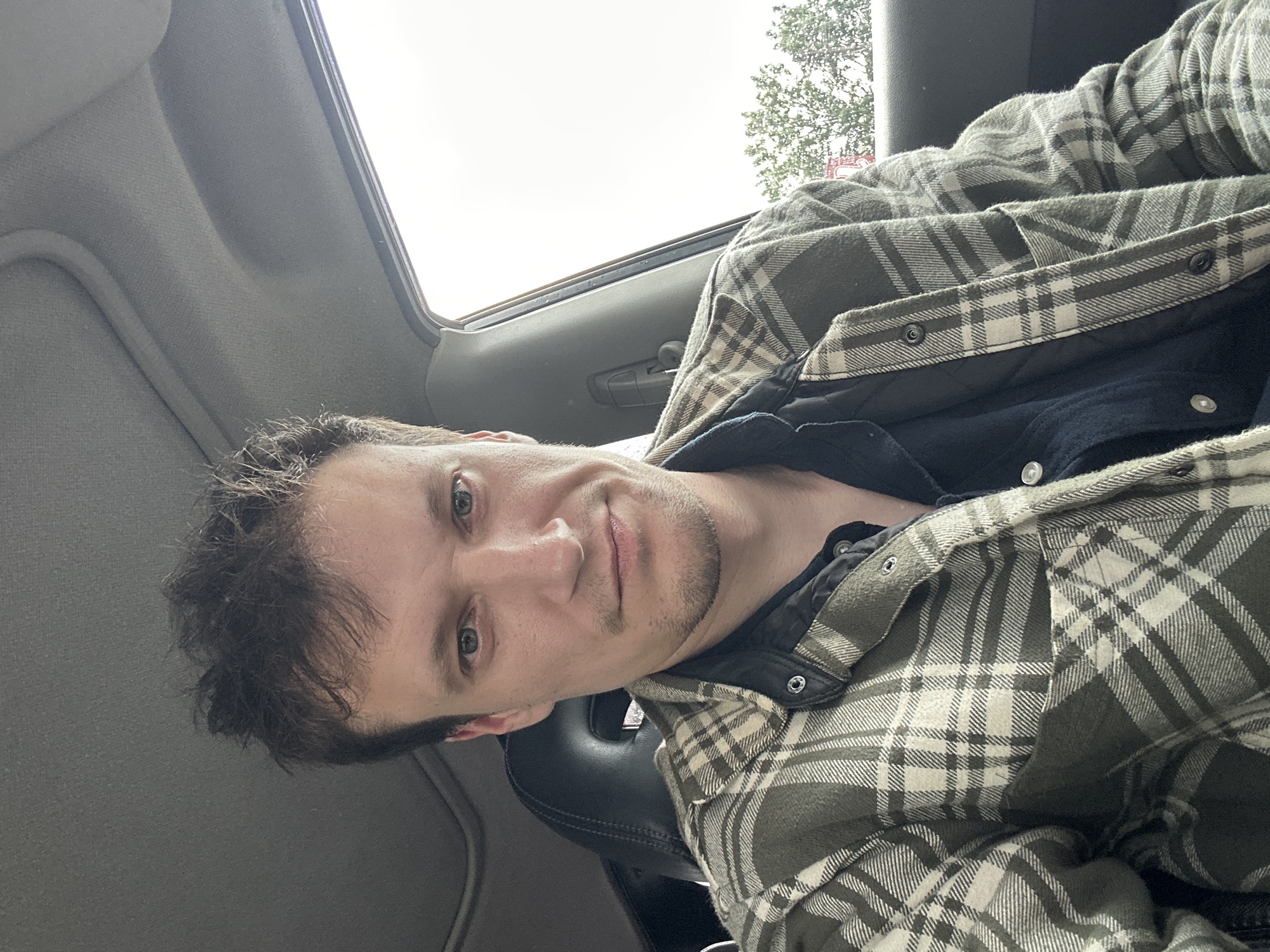 A man is sitting in the driver's seat of a car, taking a selfie. He has short, dark, messy hair and fair skin. He is wearing a dark collared shirt underneath a green and white plaid shirt jacket. The car interior is visible, including the headrest of the passenger seat and the car's ceiling. Outside the car window, green trees and a bright sky are visible, suggesting it is daytime. The man is looking directly at the camera with a neutral expression.