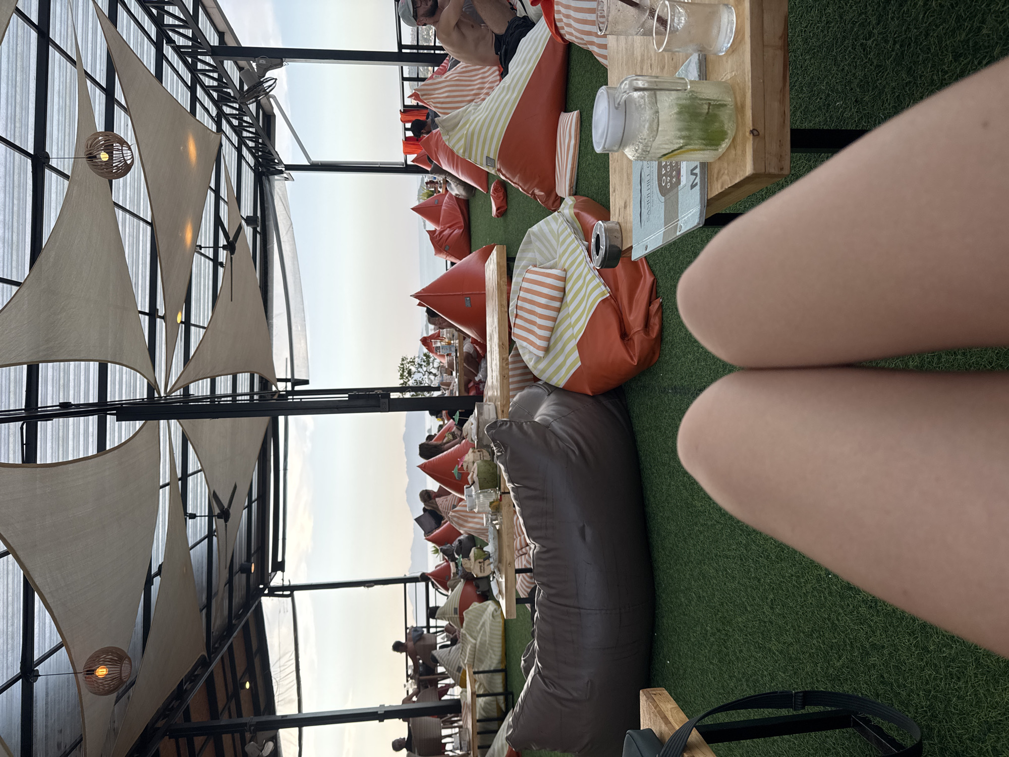 The image depicts an outdoor seating area at Samui Hill restaurant, located in Ko Samui District, Thailand. The establishment features a unique setup with an artificial turf floor and numerous beanbag chairs in orange and white stripes, as well as large, dark brown beanbags. A canopy with translucent fabric provides shade, supported by a black metal frame. Hanging lights with woven shades are visible, along with ceiling fans. In the background, a panoramic view of the ocean and distant islands is presented, suggesting a seaside location. Several patrons are seated on the beanbags, some with drinks and food on wooden tables. One table in the foreground holds a carafe of infused water with lime and mint, and a glass. The perspective is from someone seated on the floor, with their legs prominently in the foreground. The lighting suggests it is late afternoon or early evening.
