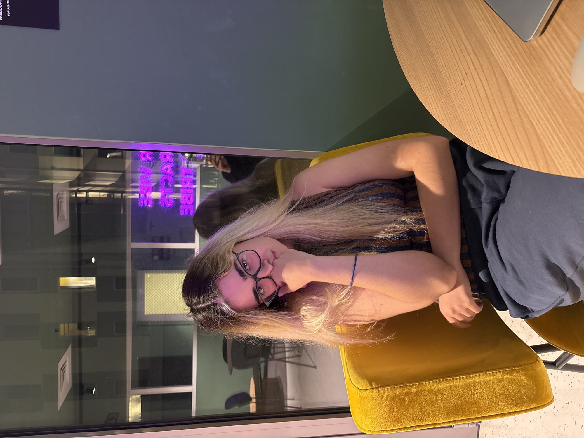A young woman with long, blonde, wavy hair and glasses is sitting in a yellow chair at a wooden table. She is wearing a striped tank top and dark blue sweatpants. Her left hand is resting on her chin, and her right arm is across her lap. She is looking directly at the camera. Behind her is a large window that reflects an interior space with purple neon lights that read "VIBE TRIBE RACERS". The floor appears to be speckled. The image is taken indoors in what appears to be a modern, possibly commercial space.