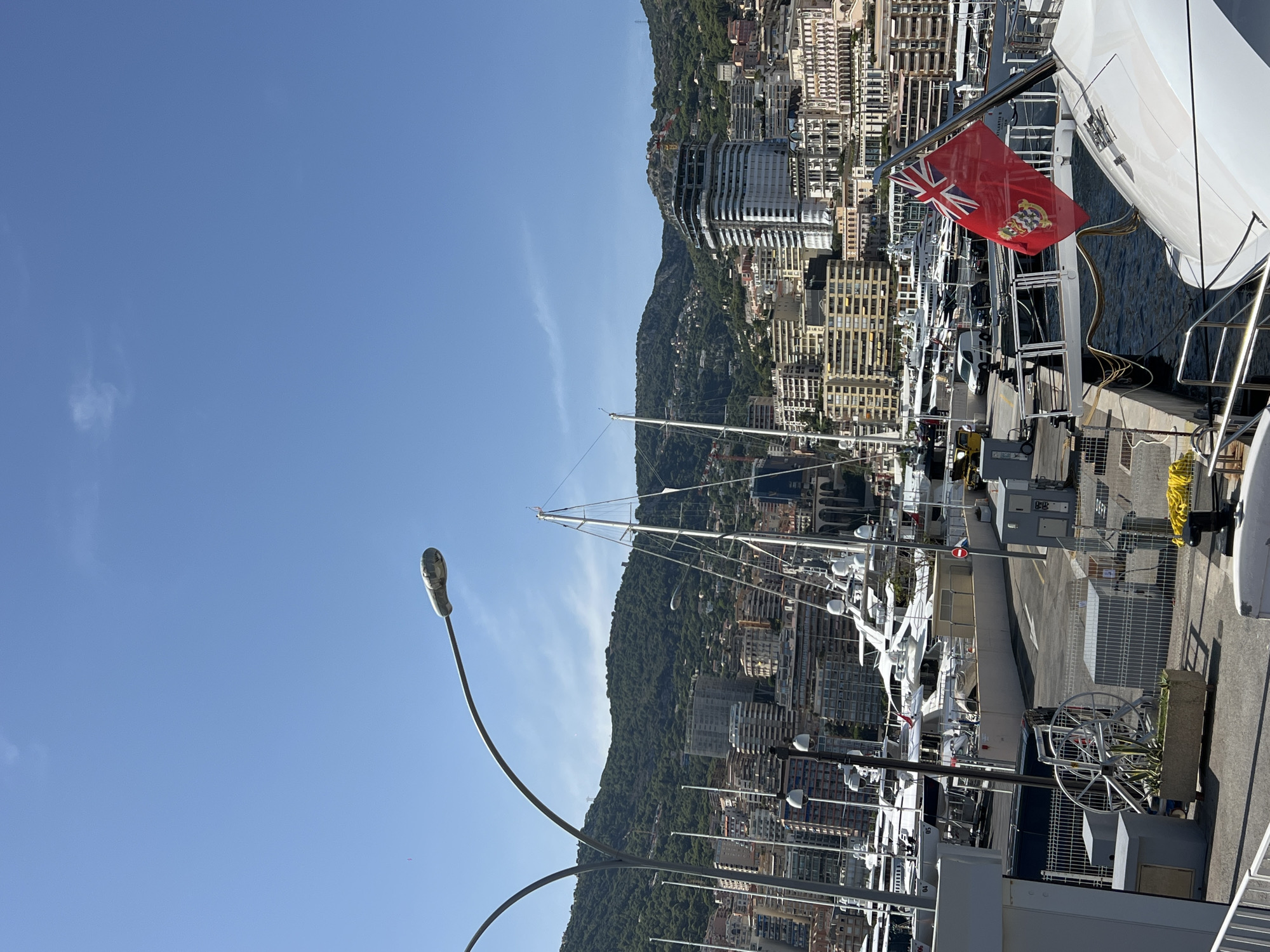 The image depicts a sunny day in the port of Monaco, likely Fontvieille, given the mountainous backdrop and the density of the cityscape. The foreground features the stern of a white luxury yacht, with a red ensign flag displaying the Union Jack and a coat of arms prominently on the right side of the frame. Mooring lines and deck equipment are visible on the yacht.
In the mid-ground, numerous other yachts of various sizes are docked in the harbor. A concrete walkway separates the yachts from a road with parked vehicles, including a white van. Several streetlights with curved arms arch over the scene. Behind the yachts, the city of Monaco rises up the hillside, characterized by a dense collection of residential and commercial buildings in shades of beige, white, and orange, interspersed with greenery.
The background is dominated by a lush, green, tree-covered mountain range that forms the natural border of the city. The sky is a clear, bright blue, indicating fair weather. A partially constructed building is visible on the hillside, suggesting ongoing development in the city. The overall scene conveys a sense of affluence and the vibrant atmosphere of a popular Mediterranean port.
