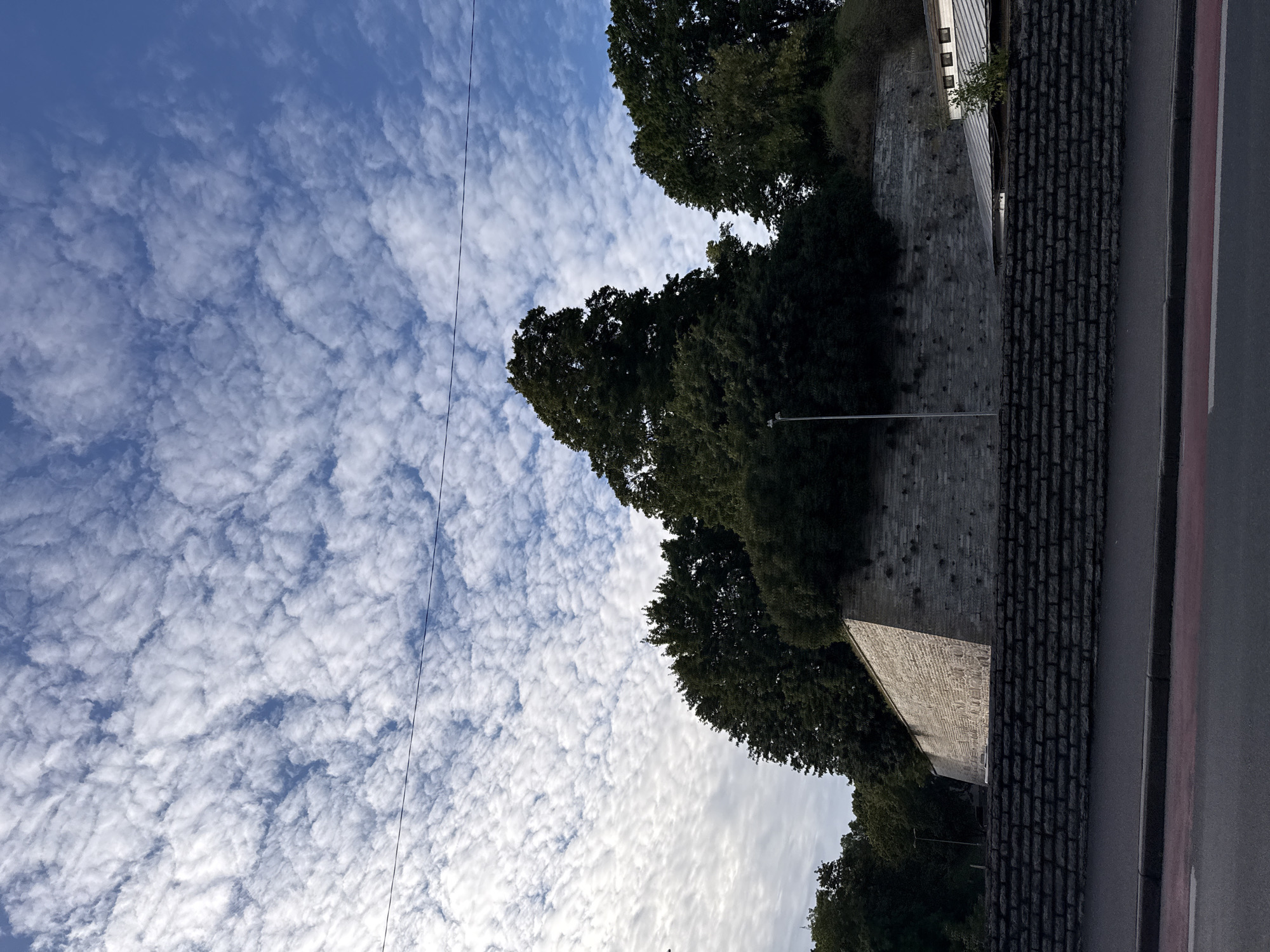A low-angle, full shot captures a stone wall and lush trees against a dynamic sky filled with cumulus clouds. The wall, constructed from large, rough-hewn stone blocks, rises from a lower, darker brick wall. The upper portion of the stone wall angles upwards, disappearing behind a dense canopy of dark green trees. The foreground features a section of asphalt road with a red-striped curb. The sky is a vibrant blue, partially obscured by a textured layer of white clouds that create a sense of depth and movement. A thin black wire stretches horizontally across the upper portion of the frame, bisecting the sky. The lighting suggests it is daytime, with shadows indicating the sun is to the left and slightly behind the viewer. The scene is devoid of people or any apparent activity.