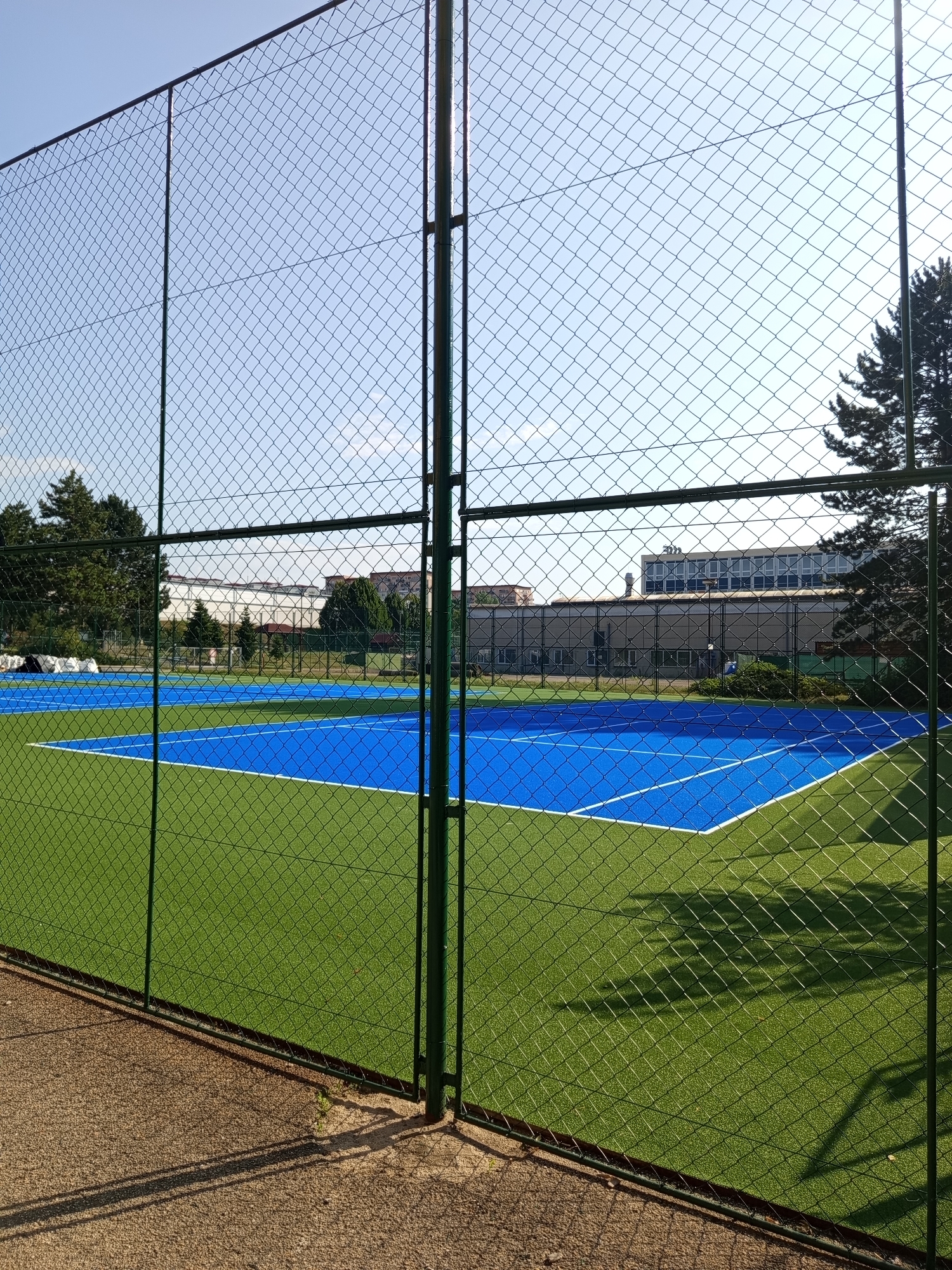 This is a photograph of a newly constructed outdoor tennis court with a vibrant blue playing surface and bright green surround, enclosed by a chain-link fence. The court is bordered by white lines. In the background, there are trees and several multi-story buildings, suggesting an urban or suburban setting. The sky is clear and blue, indicating good weather. The image was taken from outside the fenced area, looking through the fence at an angle. The shadows of the fence and possibly trees fall across the court and the surrounding area. The ground in the foreground is a gravel or decomposed granite pathway. This scene is consistent with sports facilities located within the city of Brno, Czechia, possibly in a park or recreational area. There are no people visible in the image.
