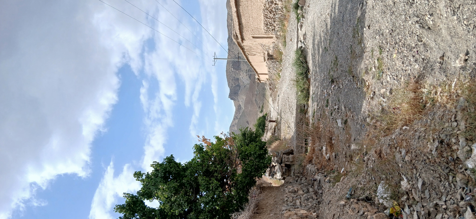 A wide-angle, vertical shot captures a desolate, dusty road winding through a rural Pakistani village, likely in Kan Mehtarzai. The sky is a pale blue, punctuated by scattered white clouds. On the left, a large, leafy green tree stands beside a low, stone-walled embankment. The embankment is sparsely vegetated with dry grass and scattered rocks. The road itself is rough and unpaved, covered in gravel and dust.
To the right of the road, traditional village homes line the path. These structures are made of mud and stone, with flat, straw-covered roofs. A utility pole with wires stretches across the upper right quadrant of the image. In the distance, rugged, arid mountains form the backdrop, their slopes barren and rocky. The overall impression is one of a remote and sparsely populated area, likely experiencing a dry climate, with evidence of poverty and limited infrastructure.