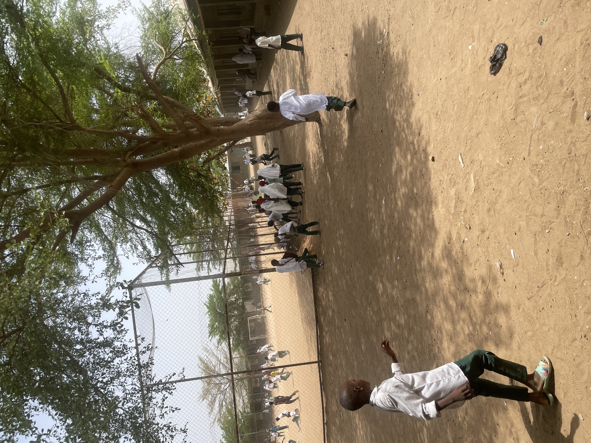 In Unguwar Rimi, Nigeria, the image shows a sandy outdoor area next to a building with several people dressed in white shirts and dark green pants. A large tree is in the foreground, casting a shadow on the ground. There is a fenced area with a soccer field visible in the background. Some people are standing in a line, while others are scattered around the field.