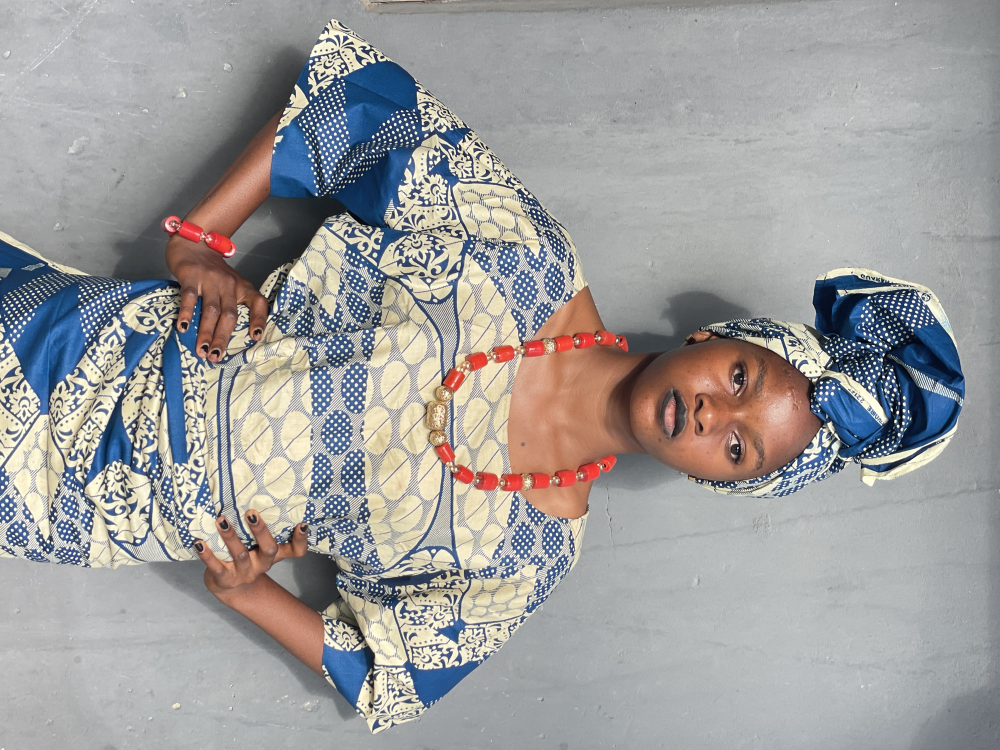 A young Black woman is standing in a gray room. She is wearing a traditional Nigerian dress with blue and off-white patterns and a matching headwrap. She also wears red beaded jewelry, a bracelet and necklace. Her hands are placed near her waist. The location is assumed to be Badagry, Nigeria.