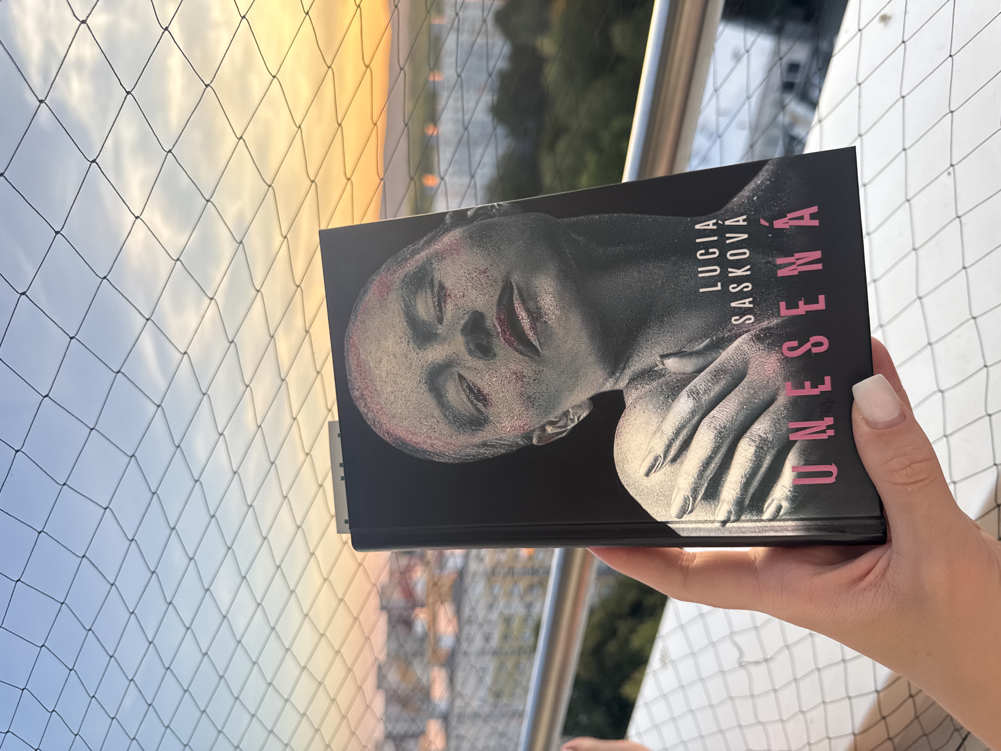 A person's hand, with white-painted nails, holds a book with a black cover featuring a close-up of a person's face with glittery makeup and metallic-looking skin. The title, "UNESENÁ," is printed in pink block letters at the bottom of the cover, above which is the author's name, "LUCIA SASKKOVÁ." The background shows a sunset over Bratislava, Slovakia, with the Danube River visible in the distance. The scene is framed by a safety net, possibly on a balcony.