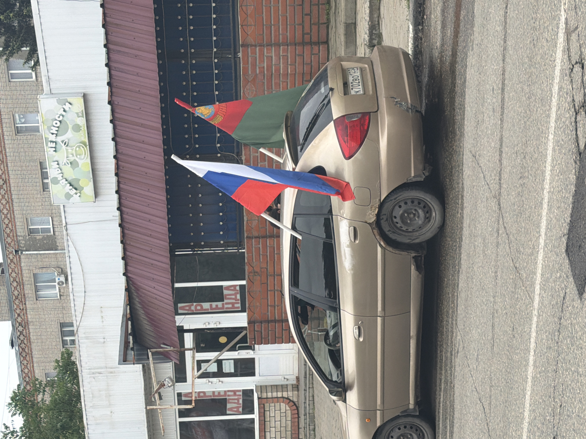 A gold-colored Kia Rio is parked on a gray asphalt road next to a building in the city of Leningradskaya, Russia. The car has visible body damage to the rear right fender. The car's license plate number is K702BO 123. The flags of Russia and Tajikistan are attached to the rear windows of the car. The building is made of brick, with a red and white awning over the storefront. There is a sign above the storefront with the word "АРЕНДА" (rent) and another sign that reads "НЕЖНОСТИ ТЕЛЯЧЬИ МЯСА". The day is overcast and lighting is diffused.