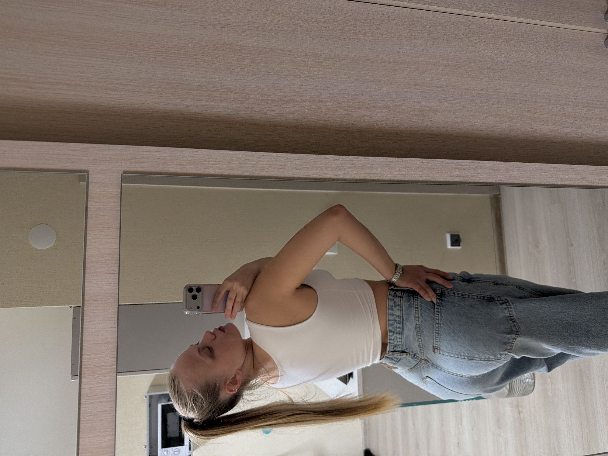 A woman is standing in front of a mirror taking a selfie with a phone. She is wearing a white tank top and blue jeans. Her hair is pulled back in a ponytail. The background includes a light-colored wall and a mirror reflecting the scene. The reflection suggests a bathroom setting with a cabinet and potentially a small appliance like a microwave visible. The location is Izhevsk, Russia.