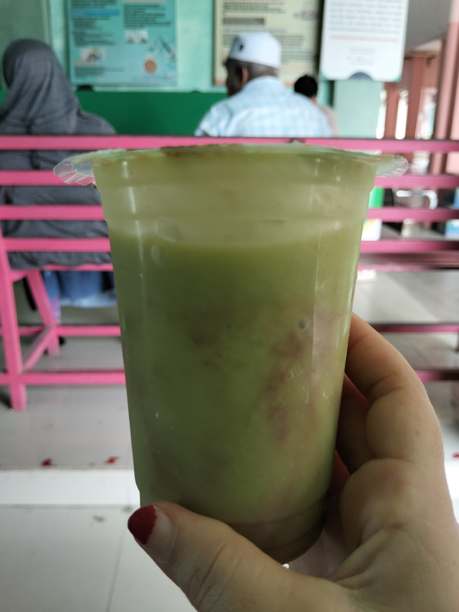 A close-up shot focuses on a clear plastic cup filled with a pale green, frothy beverage, likely a smoothie or juice. The drink has some darker, reddish-brown streaks mixed within it. The cup is held by a hand with neatly painted red fingernails.
In the blurred background, a person wearing a grey hijab is visible on the left, seated on a pink bench. To the right, another person wearing a white skullcap and a blue and white checkered shirt is also seated. The setting appears to be a casual indoor or semi-outdoor eatery, possibly a food stall or small restaurant, indicated by the colorful benches and posters on the wall. The poster on the left wall has some green and blue hues with visible text and graphics, but it is too blurred to decipher. The poster on the right is mostly white with some red text. The overall lighting suggests it is daytime. The mood appears relaxed and casual. The location context provided is Bireuen, Indonesia.