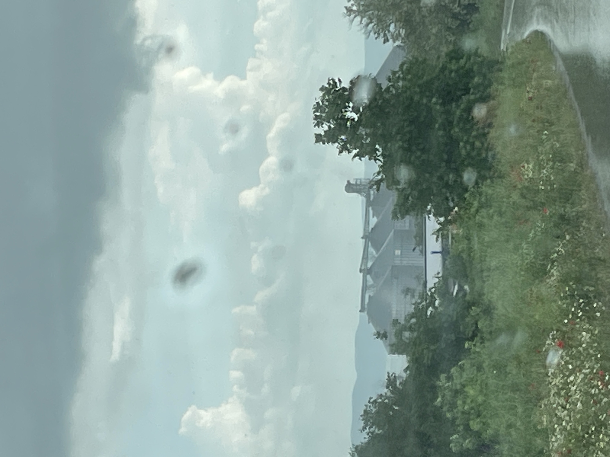 The image depicts a cloudy sky with a visible industrial structure in the background, likely a storage facility, possibly a grain elevator. There are trees and greenery along what appears to be a road or path on the right side of the image, with some red flowers interspersed. The presence of water droplets suggests rain or a wet surface. The photo was taken in Makariopolsko, Bulgaria.