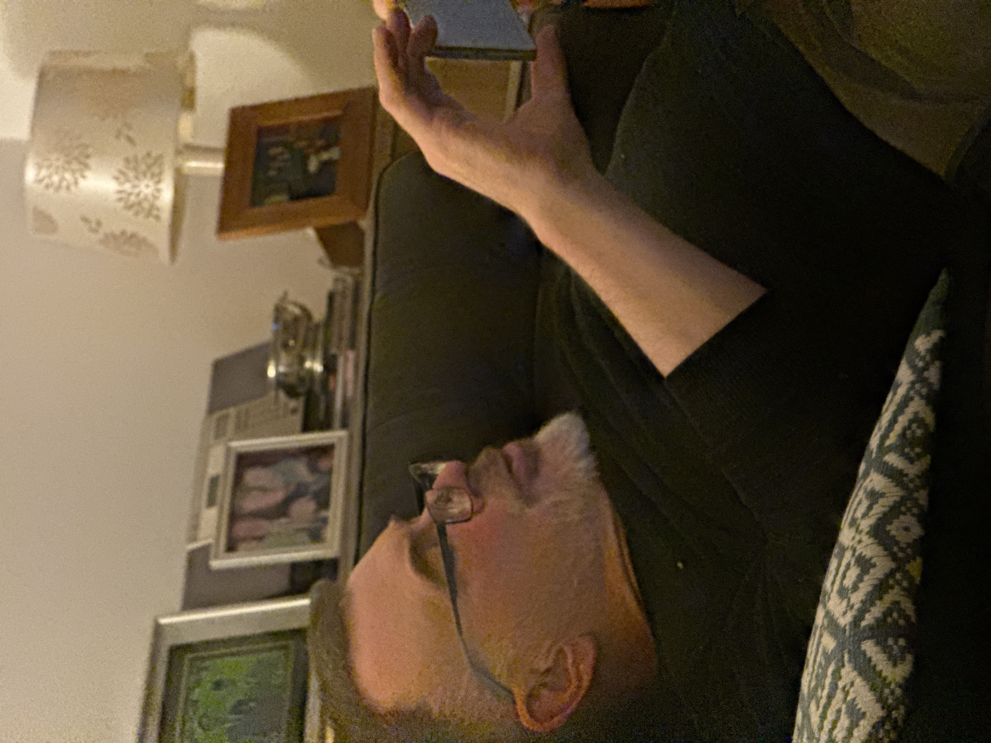 An adult male with glasses, fair skin, and short grey facial hair is reclined, facing left, holding a dark object in his right hand near his head. He is wearing a dark shirt and appears to be resting against a patterned pillow on a sofa. Behind him and to the left is a wall featuring a lamp with a patterned shade and several framed photographs. The location is Unknown, Unknown.