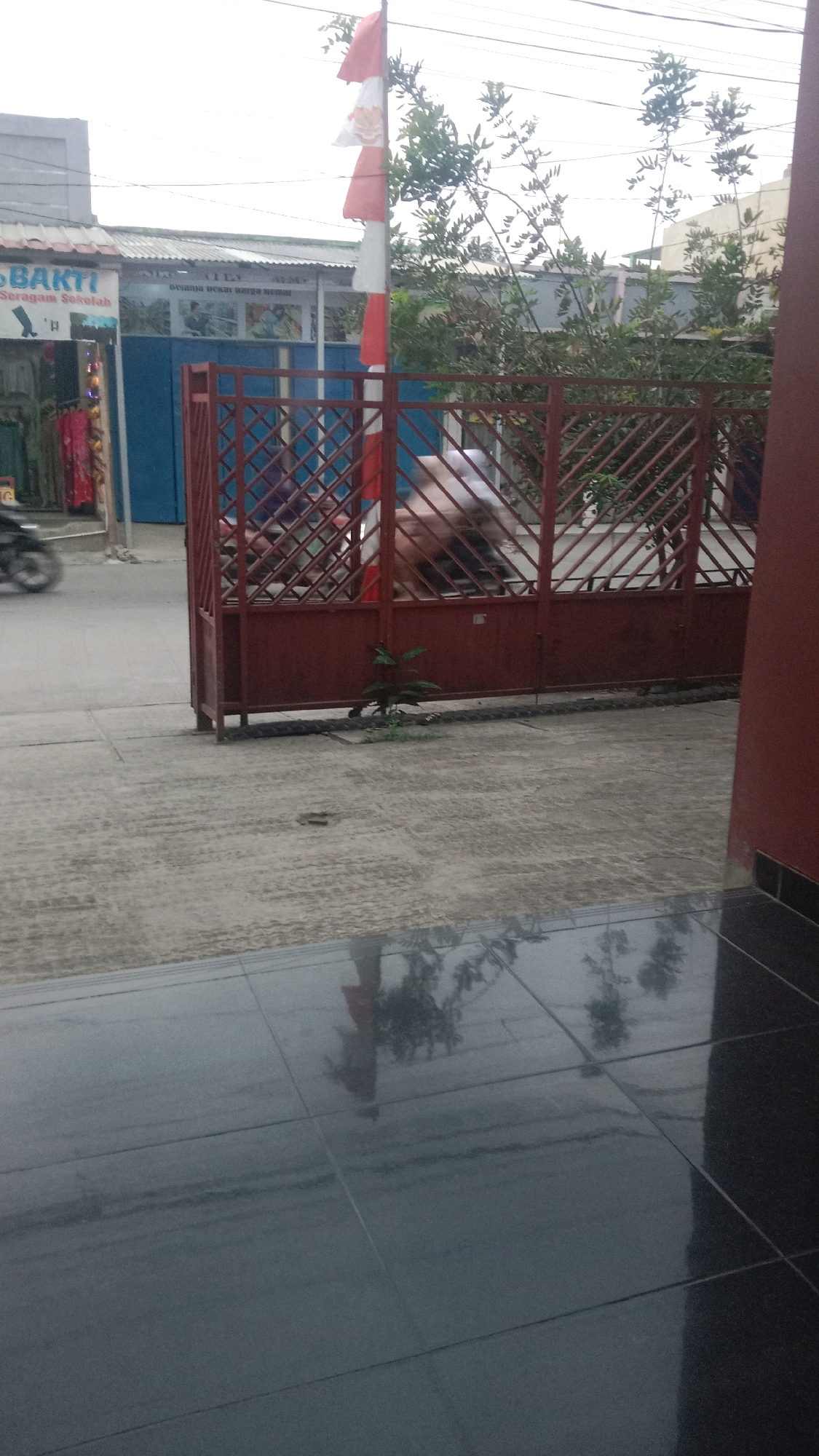 In the city of Jalenjaya, Indonesia, an outdoor scene unfolds with a red metal gate as a central element. The gate, with its diamond pattern, is partially open, allowing a glimpse of movement within. To the left of the gate, a store with the text "BAKTI" and "Seragam Sekolah" is visible, displaying various school uniforms. A red and white Indonesian flag is prominently displayed on a flagpole to the left of the gate, its colors stark against the overcast sky. Behind the gate, a blurred figure on a motorcycle can be seen, suggesting traffic or movement beyond the immediate foreground. The ground is paved with concrete, marked with tire tracks and a few small plants. The reflection of trees and possibly the flag can be seen on the dark, glossy tiled surface in the foreground, indicating a polished floor or a wet surface. The overall environment appears to be a street or pathway in a commercial or educational area.