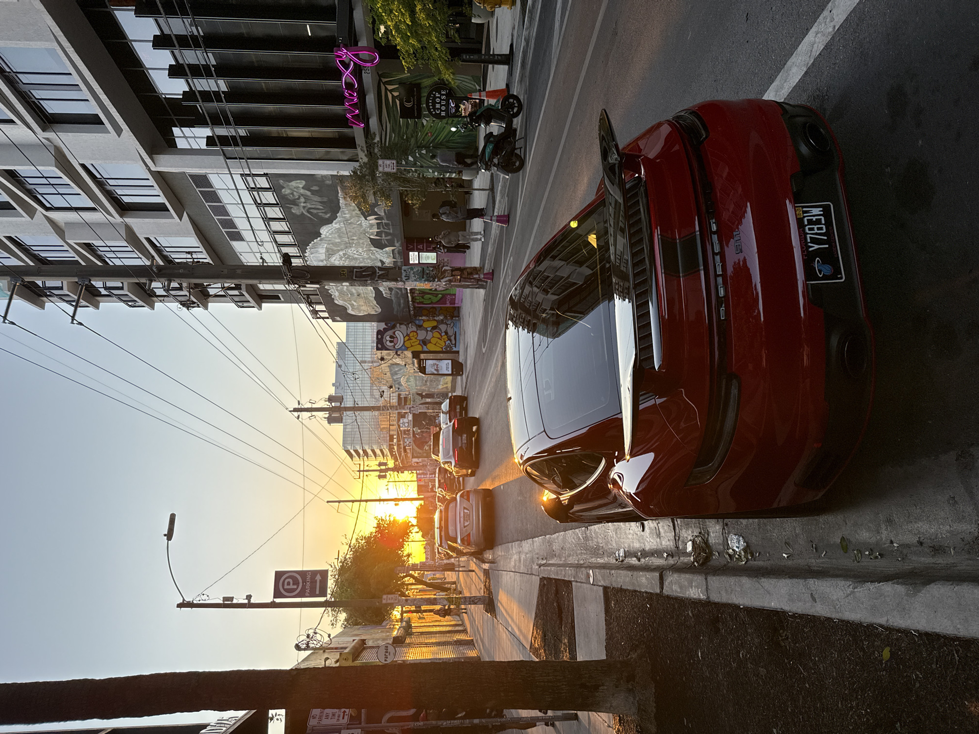 An urban street scene in the Wynwood Art District of Miami, Florida, USA, is depicted during golden hour, characterized by warm, low-angle sunlight from the west.
In the immediate foreground, positioned on the right, a red Porsche 911 (992 generation), identifiable by its rear design, black rear spoiler with a red accent, and dual exhaust outlets, is parked parallel to a concrete curb. The vehicle bears a Florida license plate, "Y783W," mounted in a "Kaseya Center" frame displaying the Miami Heat logo. The asphalt street surface features a white parking line adjacent to the Porsche. The curb line exhibits minor debris and scattered leaves.
On the right side of the street, a modern multi-story building, identified by a prominent pink neon "moxy" sign and a smaller "255" address marker, houses the Moxy Miami Wynwood hotel. Below the Moxy sign, a planter box displays a "BROOKLYN CHOP HOUSE ROOFTOP SOCIAL CLUB" sign. Adjacent to this structure, a wall features a large black-and-white mural depicting an alligator or crocodile. Further left, another building exhibits vibrant, cartoonish graffiti art, including a yellow character face. On the sidewalk, a turquoise motor scooter is parked angled, and three adult individuals are standing or conversing. Two orange traffic cones are visible on the sidewalk near the scooter, and a yellow fire hydrant is positioned further down the street.
On the left side of the street, a tree trunk is present in the foreground. A concrete sidewalk parallels the road, where one pedestrian is visible walking away from the camera, illuminated by the sun. Multiple "NO PARKING ANY TIME" signs and a "P" parking sign with an arrow indicating parking direction are mounted on utility poles along the sidewalk.
The roadway, leading into the distance, contains several vehicles in traffic. A prominent silver sedan is visible, reflecting the sunlight, followed by a dark-colored Porsche and other automobiles.
The background shows a low sun on the horizon, casting an orange-yellow glow that transitions to a light blue sky overhead. Numerous overhead power lines crisscross the frame. Distant city buildings are partially visible, reflecting the ambient light. The scene is illuminated by strong, warm sunlight, creating long shadows and prominent reflections on surfaces.
