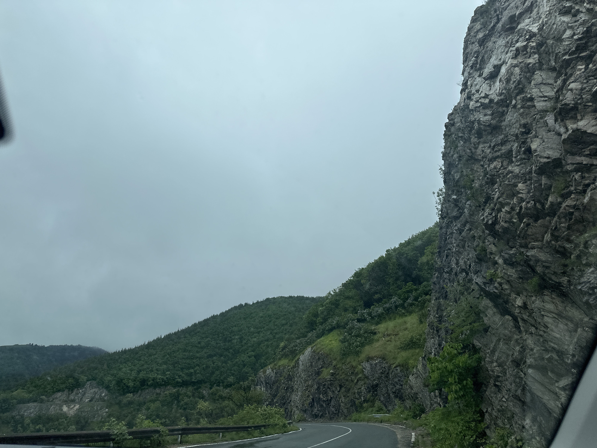 The image depicts a winding road in Karnare, Bulgaria, cutting through a mountainous landscape under a cloudy sky. A steep, rocky cliff face dominates the right side of the frame, while a forested hillside extends to the left. A guardrail runs along the edge of the road. The road surface is wet, suggesting recent rain. The scene appears to be viewed from inside a vehicle. No people or animals are visible.