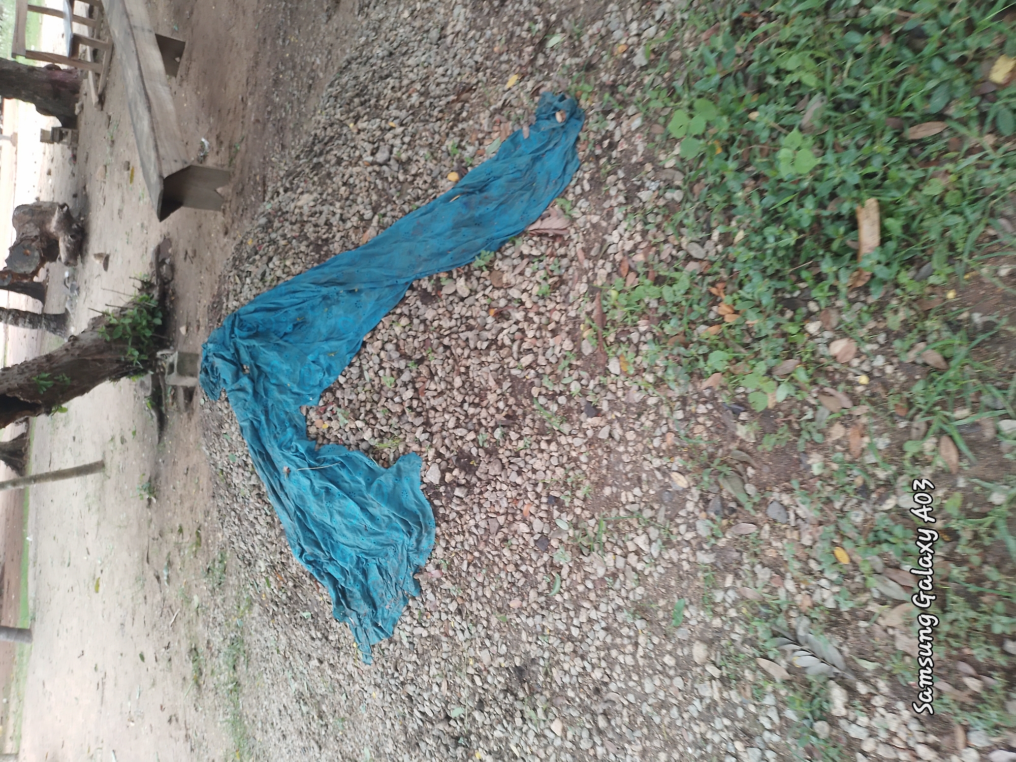 A blue tarp is draped over a pile of gravel. The pile of gravel is located outdoors, with trees and dirt ground surrounding it. In the background, a wooden bench is visible, along with more trees and some fallen logs. The scene suggests an outdoor construction or storage area in Doka-Tofa, Nigeria.