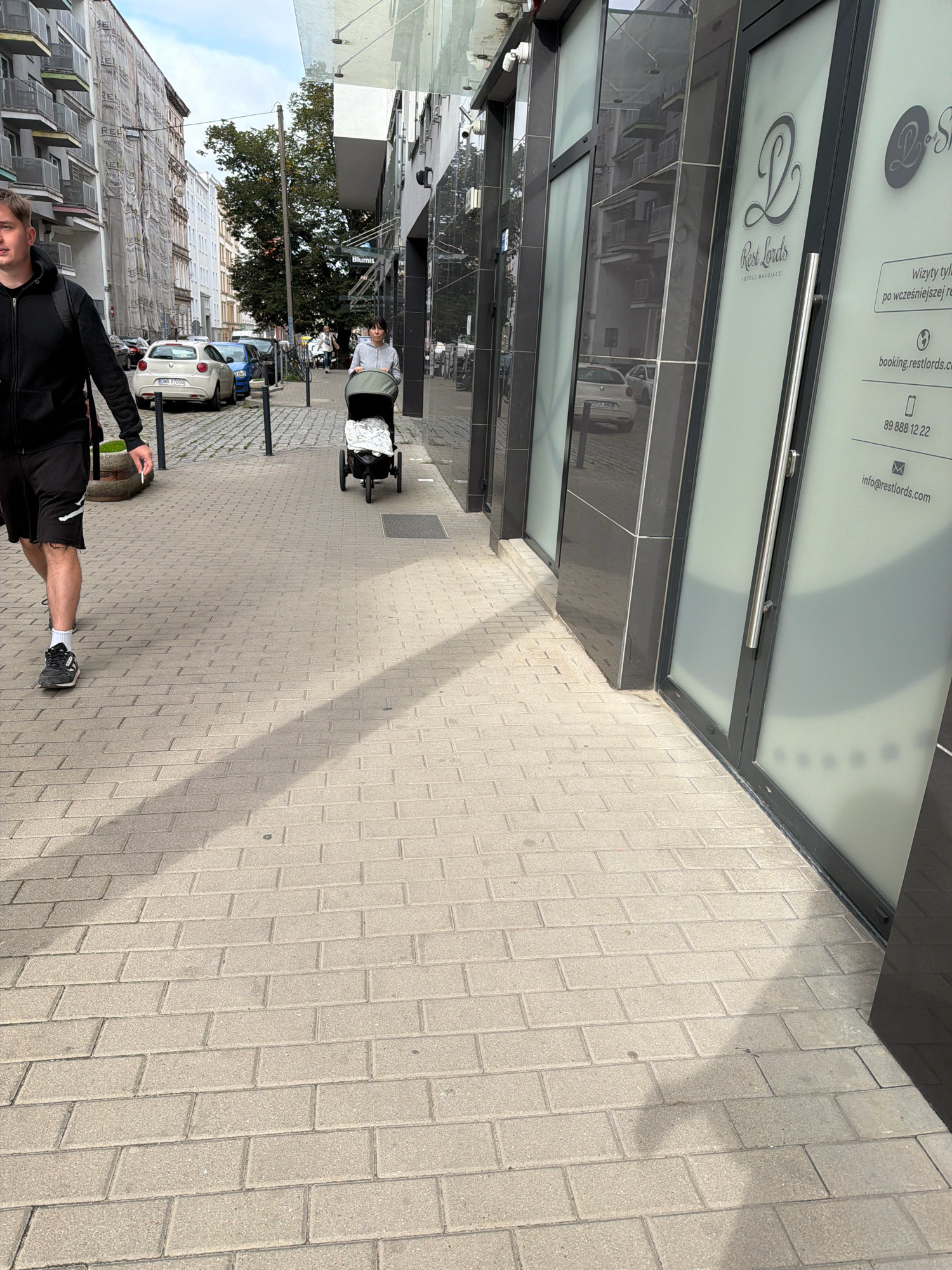 The image depicts a sidewalk in Wrocław, Poland, on a cloudy day. A man wearing a black hoodie and shorts walks towards the camera, holding a cigarette. Behind him, a woman pushes a stroller. On the right side of the image, a building with frosted glass doors displays advertisements for "Rest Lords" and contact information. Cars are parked along the street, and buildings with balconies are visible in the background. The sidewalk is paved with light-colored rectangular bricks.