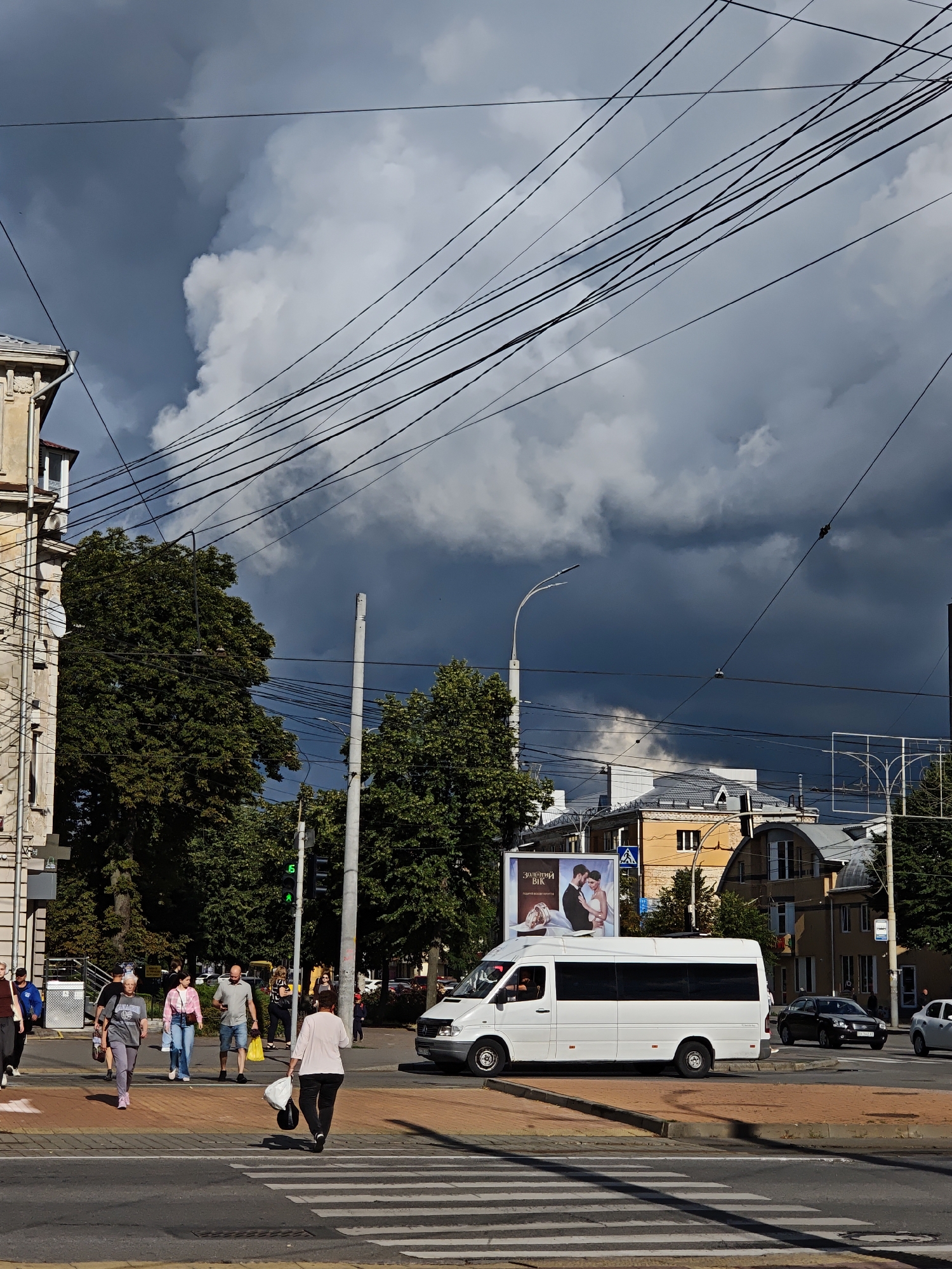 A street scene unfolds under a dramatic, cloudy sky in Vinnytsia, Ukraine. The time of day appears to be late afternoon or early evening, as evidenced by the strong shadows. Dark, heavy clouds dominate the upper portion of the frame, suggesting an approaching storm, contrasting with patches of brighter sky peeking through.
In the foreground, a pedestrian crossing is occupied by several individuals. A woman in a light pink top and black pants walks away from the viewer, carrying shopping bags. Behind her, a woman in a grey t-shirt and light grey pants walks towards the crossing, also with bags. A couple, a man in a grey polo shirt and shorts, and a woman in a pink jacket and jeans, are also crossing. Another person in a dark top and grey pants walks towards the crossing. A white minibus is stopped at the crossing, its windows tinted dark. Beyond the minibus, a black sedan and a white car are visible on the road.
The setting is urban, with buildings lining the street. To the left, a light-colored building with ornate architectural details stands tall. Trees with lush green foliage are abundant, framing the scene and providing shade. Overhead, a complex network of utility wires crisscrosses the sky. A billboard is positioned behind the minibus, displaying an image of a couple and text in Ukrainian which includes "Золотий Вік" (Zolotyy Vik), meaning "Golden Age". A traffic light with a green signal is visible on a pole to the right of the crossing. The overall mood is one of everyday life transitioning under the impending weather.