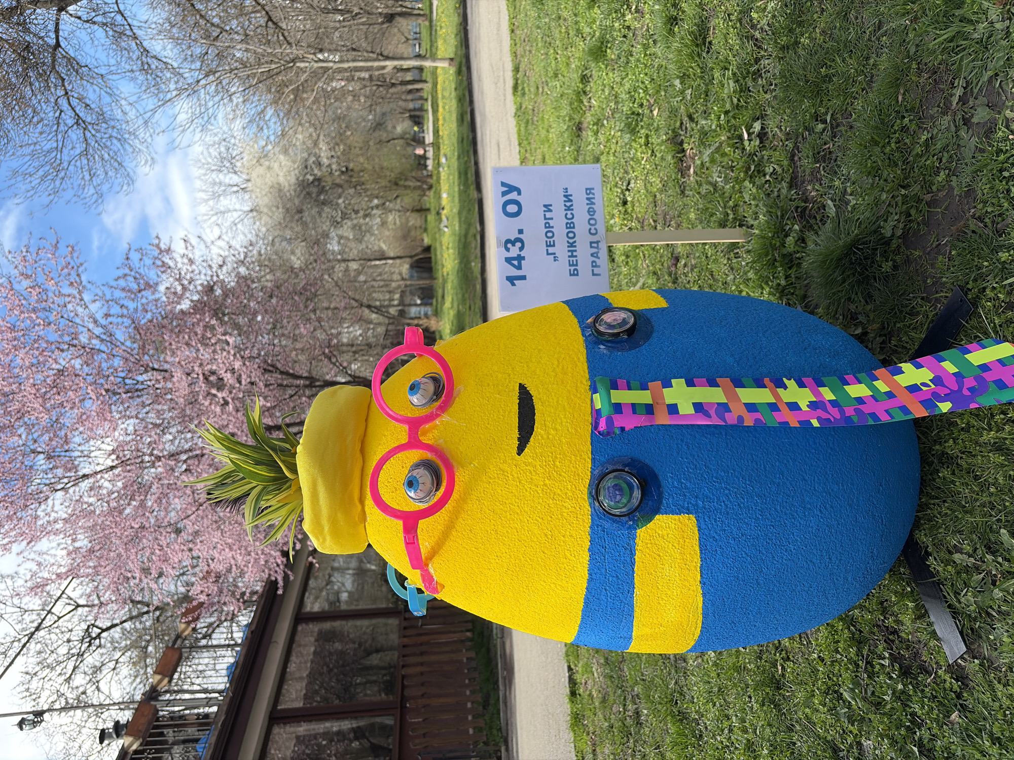 A low-angle shot depicts a whimsical, Minion-like figure in an outdoor setting. The figure, composed of textured yellow and blue sections, is adorned with oversized pink glasses, a yellow beanie, and a tuft of green pineapple-like leaves on its head. Its body features what appear to be protruding eyes and a simplistic black smile. A colorful, patterned ribbon is affixed to the side of the figure.
Behind the figure stands a sign with text written in Bulgarian, indicating "143. ОУ 'Георги Бенковски' Град София," suggesting that this display is located near or associated with the 143rd school named after Georgi Benkovski in the city of Sofia, Bulgaria.
The backdrop includes green grass, budding pink-blossomed trees, a building with windows, and a blue sky with scattered clouds. The scene suggests a public space such as a park or schoolyard.
