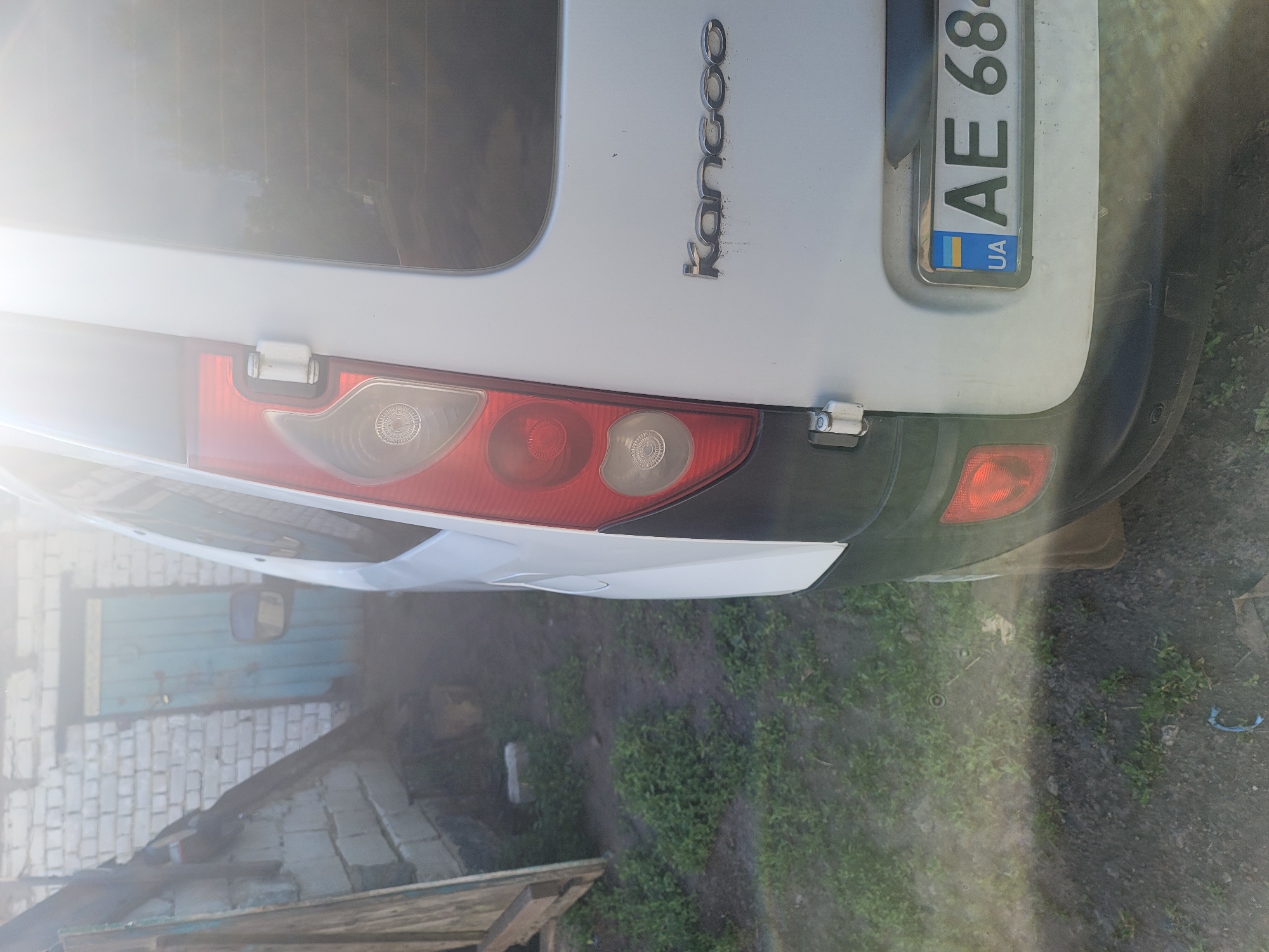In Vovkivka, Ukraine, a white Renault Kangoo van is parked outdoors, with its rear visible. The van has a vertically oriented taillight assembly on the left side, featuring red and clear elements. A chrome "Kangoo" emblem is affixed to the right side of the tailgate. A license plate, with the identifier "AE 684" and a Ukrainian flag emblem, is mounted on the rear bumper. The van is positioned on a dirt and grass surface, with a weathered brick building and a blue wooden door visible in the background to the left. The scene is illuminated by bright sunlight, causing lens flare and overexposure in some areas of the image.