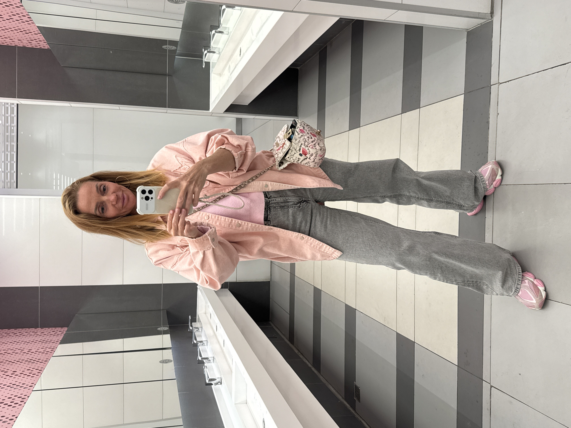 A woman stands in a bathroom, taking a selfie in the mirror. The woman is wearing a pink jacket, pink sweater, gray jeans, and pink sneakers. She has long blonde hair. She is holding a white cell phone. Her small white purse is on her left shoulder. The bathroom has a long white sink with silver fixtures, and the floor is tiled in a gray and white pattern. The walls are white with gray accents. There is a pink patterned design on the left-hand wall. There is no indication in the image that identifies the location as Saint Petersburg, Russia.
