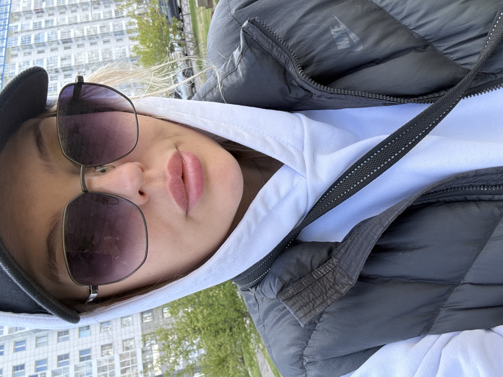 A woman with blonde hair is wearing sunglasses and a cap. She is wearing a white hooded sweatshirt under a black Nike puffer vest. A black strap runs diagonally across her body. The background shows an apartment building and trees, likely in Krasnoyarsk, Russia.