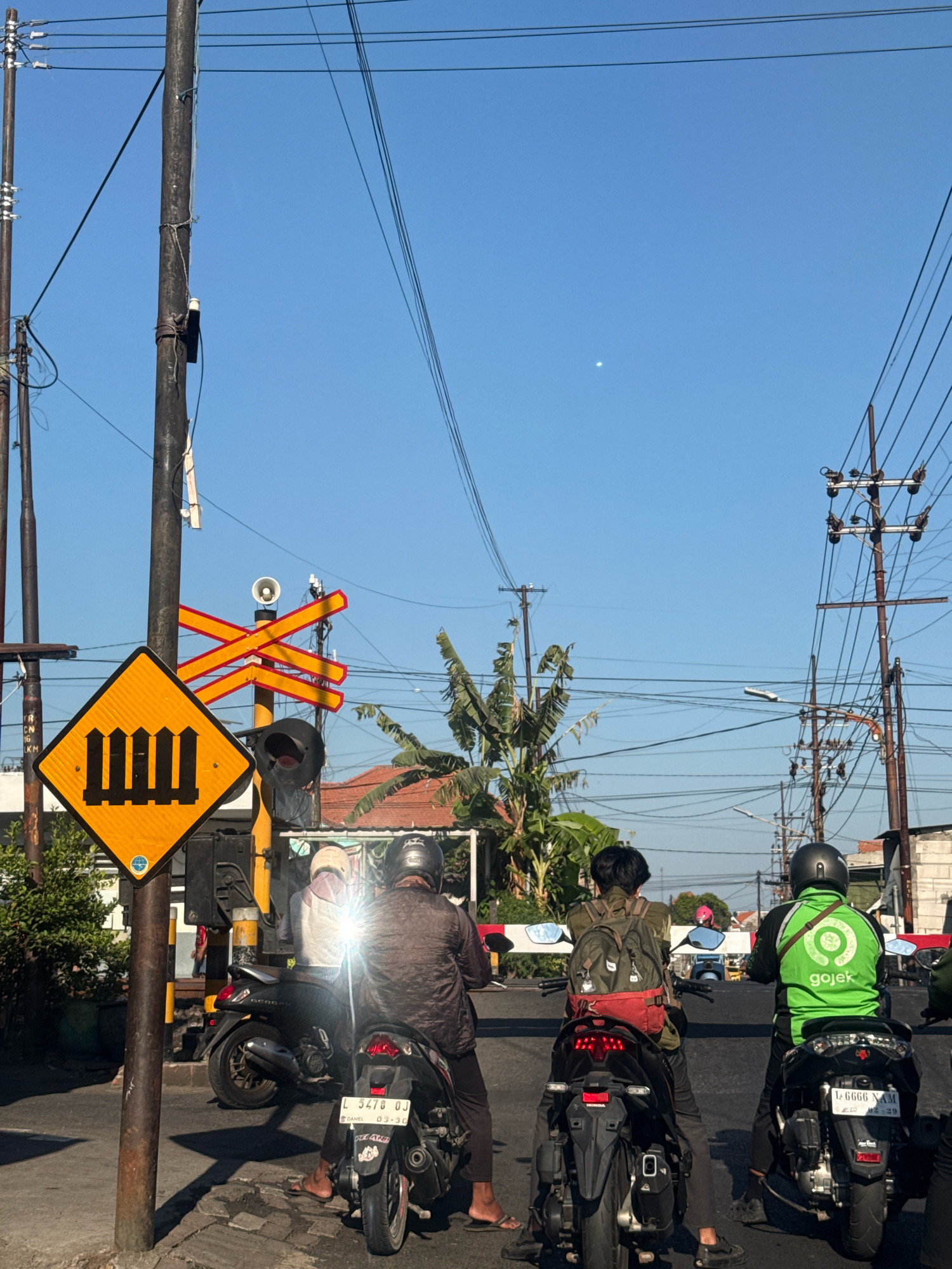 In Surabaya, Indonesia, a street scene unfolds under a clear blue sky, featuring a railroad crossing with barriers, traffic lights, and utility poles adorned with numerous wires. At the crossing, three individuals on motorcycles are visible. One motorcycle, a black scooter, is ridden by a person wearing a brown jacket and a helmet, with another person behind them wearing a white headscarf and a light-colored vest. Another black scooter is ridden by a person with a backpack, and a third black scooter is ridden by a person in a bright green jacket with the "Gojek" logo, indicating a ride-sharing service. The ground is paved with a combination of cobblestones and asphalt. In the background, lush green trees and buildings suggest a suburban or semi-urban environment. The image captures a moment of waiting at the railroad crossing, likely for a train to pass.