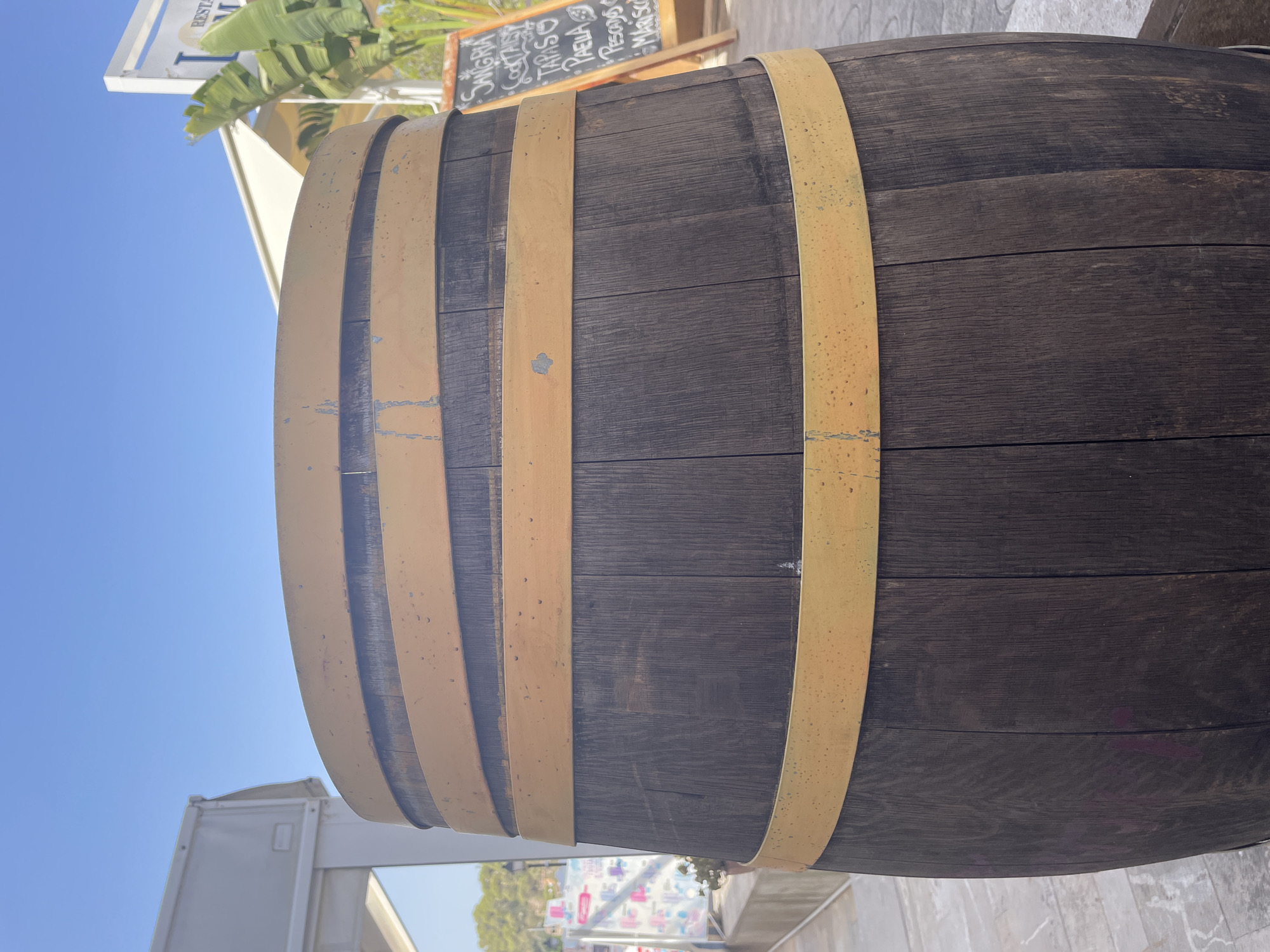 The image shows a close-up, head-on view of a wooden barrel with yellow bands, situated outdoors in what appears to be a commercial area. The barrel is the primary subject, dominating the foreground. Behind it, out of focus, are elements of a street or promenade with paved flooring and a building with a white awning or roof. To the right of the barrel, a small chalkboard menu is visible, leaning against a stand, displaying handwritten text that includes "SANGRIA," "COCKTAILS," "TAPAS," "PAELLA," "PESCADO," and "MARISCO." A palm frond is also visible in front of the chalkboard. The sky is clear blue. The overall scene suggests an outdoor seating area of a restaurant or bar in a Mediterranean setting. The location is identified as Santanyí, Spain. There are no people visible in the frame.