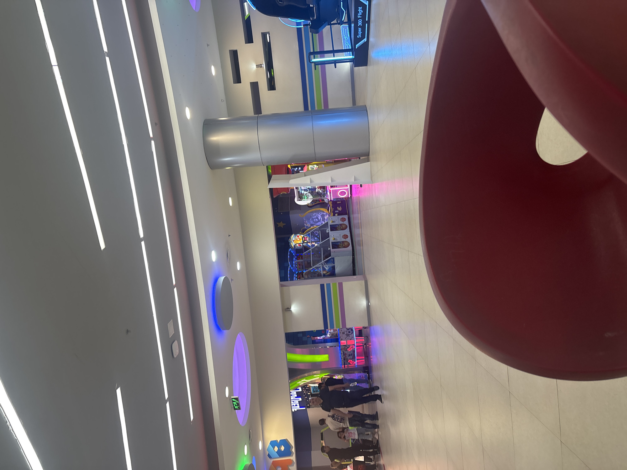 The image depicts an indoor entertainment area, likely an arcade or a section within a shopping mall, in Yerevan, Armenia. The scene is brightly lit with numerous artificial light sources, including long fluorescent strips on the ceiling and walls, and circular/oval light fixtures emitting blue, purple, and green hues.
A prominent feature in the midground is a large, reflective silver cylindrical column. In the background, a variety of colorful arcade games and amusement rides are visible, characterized by their illuminated screens and vibrant designs. One game is clearly labeled "Super 360 Flight."
On the far right, a large, curved red sculptural element or piece of furniture dominates the foreground, partially framing the scene. Several people, including adults and children, are gathered and walking in the lower right portion of the image, near the arcade games. They are casually dressed, engaged in typical activities for such a venue.
The flooring is a light-colored tiled surface. A green exit sign with an arrow is visible on a wall. The overall ambiance is lively and recreational, consistent with an entertainment zone, with the bright artificial lighting creating an atmosphere that could be at any time of day, but often suggests evening or night in such venues.