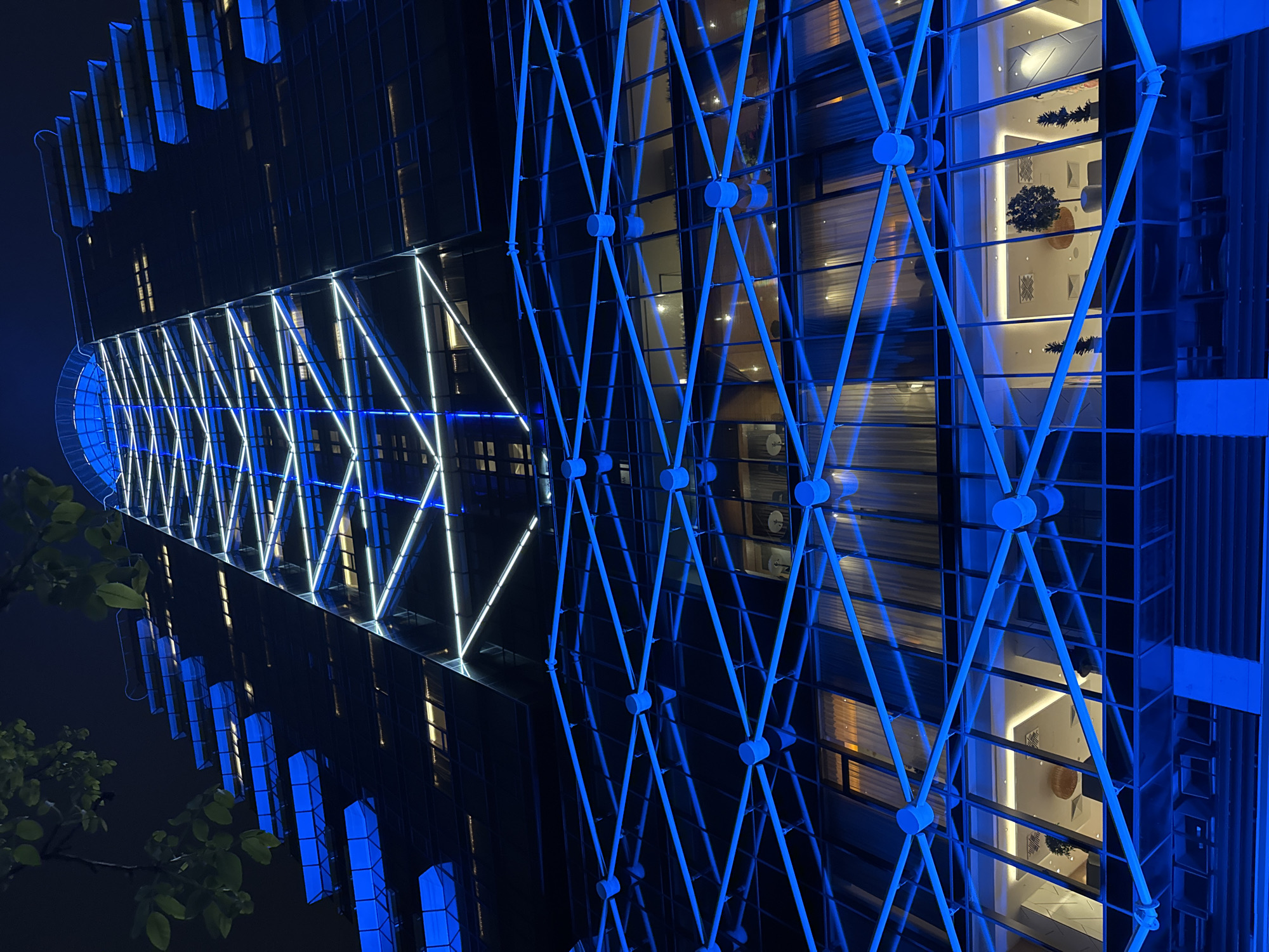 The image shows a tall building at night, likely in Sofia, Bulgaria, given the prompt's request. The building is characterized by a modern design featuring a geometric facade, illuminated with white and blue lighting. The building's glass panels reflect the internal lighting. A diamond-shaped steel structure overlays parts of the facade. The environment is dark. There are leaves from a nearby tree visible in the lower left corner. The building's interior, glimpsed through the glass, reveals decorated spaces with plants.