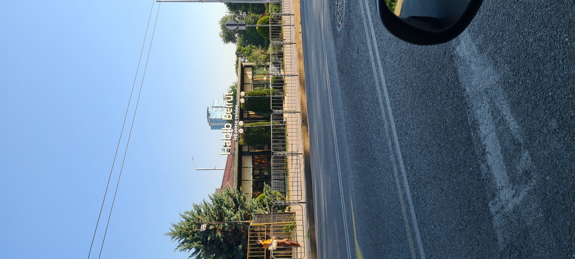 A low-angle shot captures a street scene in Sofia, Bulgaria, on a clear day. In the foreground, the asphalt road with white lane markings dominates the lower half of the frame. A car's side mirror is partially visible on the right. A metal fence separates the road from a sidewalk and a building.
The building, identified by signage as "Habib Beirut Lebanese cuisine," features a large sign with illuminated white lettering. It has a brown roof and large glass windows, with manicured bushes and trees adorning its facade. A woman wearing a yellow top and brown pants stands near a gate to the left of the building.
Behind the restaurant, a modern glass skyscraper stands tall against the blue sky. Trees and utility poles are also visible in the background. The lighting suggests it is daytime, likely late afternoon, given the shadows.