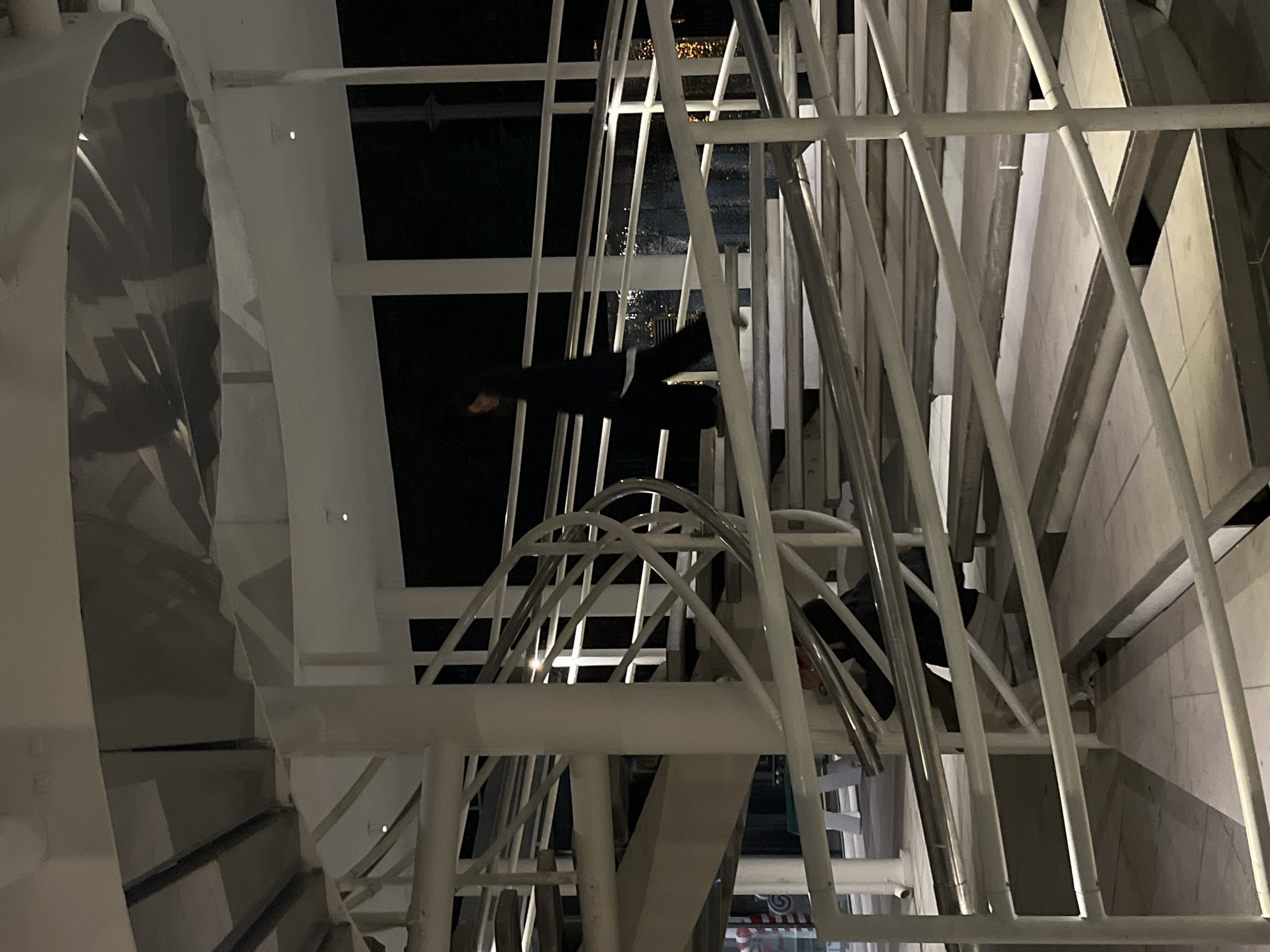The image captures a nighttime scene, likely in Moscow, Russia, dominated by a complex, white architectural structure. The primary subject is a person, appearing to be an adult, dressed in dark clothing (possibly a black jacket or coat), standing or leaning within the intricate framework of this structure. Their face is obscured by shadows.
The structure itself consists of numerous white, slender metal bars, forming a grid-like or lattice pattern, suggesting it could be part of a large spiral staircase, a decorative facade, or an observation wheel. To the left, a large, dark, ribbed or fluted cylindrical object is visible, appearing to be part of the building or a significant architectural feature. The overall design features both straight and curved elements, creating a dynamic and geometric composition.
The background is dark, indicating it is nighttime. Distant, blurry lights are reflected in what appears to be a wet or highly reflective surface below, possibly water or a polished ground, hinting at urban surroundings. The interplay of light and shadow highlights the three-dimensional nature of the white structure. The image shows no clear activity beyond the person's presence. There is a very small, unreadable red and white logo or text partially visible at the bottom right corner of the original image.