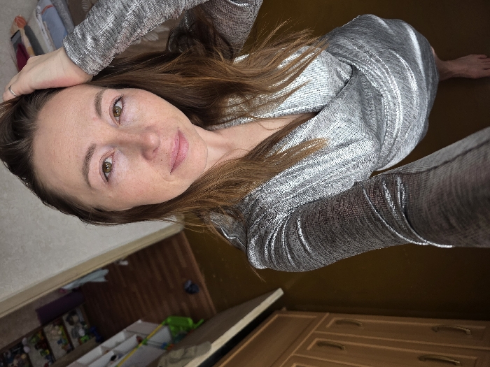 The image depicts a high-angle self-portrait of a female individual within an indoor residential setting, reportedly located in the city of Chaikovsky, Russia. The subject, an adult female with medium-length light brown hair and a fair complexion, looks directly into the camera with a neutral expression. Her eyes are greenish-brown, and she has visible freckles on her nose and cheeks. She is wearing a shimmering silver or metallic-grey long-sleeved dress or top featuring a wrap-style V-neckline. Her left hand is raised, with fingers lightly touching her hair near the left temple; a silver-colored ring is visible on her left ring finger. Her bare feet are visible at the bottom of the frame.
The immediate environment features a dark brown floor, possibly wood or laminate. To the left, a light brown wooden cabinet with brass-colored handles is partially visible. Beyond the cabinet, an open doorway reveals an adjacent room with a lighter brown wooden floor. Within this secondary room, white shelving units against a light wall are filled with various small objects, including what appears to be children's toys and containers. A green plastic item rests on the floor near the doorway, and a dark shoe or object is further into the room. To the upper right of the subject, a light-colored wall is visible, along with a stack of folded textiles (red, white, and patterned) on an unseen surface. The lighting appears to be interior ambient light, highlighting the texture of the subject's attire.