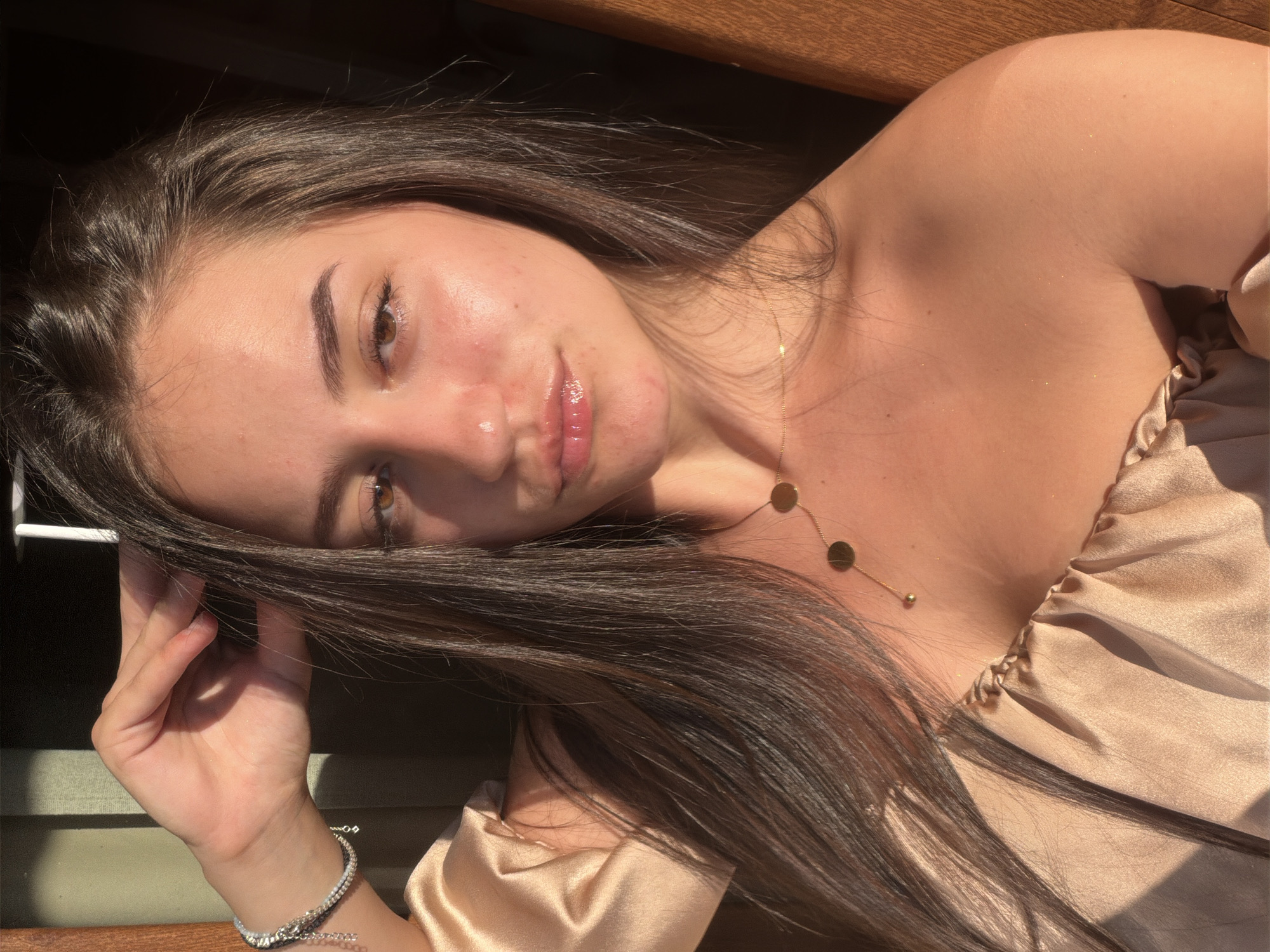 The image is a close-up, angled selfie of a young woman. She has long, dark brown hair that partially obscures her face, and is looking towards the camera. Her skin appears sun-kissed and has a subtle sheen, with a few small blemishes visible on her cheeks. She is wearing a light, off-the-shoulder top made of a satin-like fabric in a champagne or beige color. A delicate gold necklace with circular pendants rests on her collarbone. On her left wrist, she wears a sparkling bracelet. The lighting suggests it is daytime, with strong sunlight casting shadows and highlighting parts of her skin and hair. The background appears to be a wooden surface, possibly a bench or table, with some darker shadows suggesting an outdoor or semi-outdoor setting. The overall mood conveyed is relaxed and natural, with a touch of casual elegance. The image was taken in Burgas, Bulgaria. There is no visible text in the image.