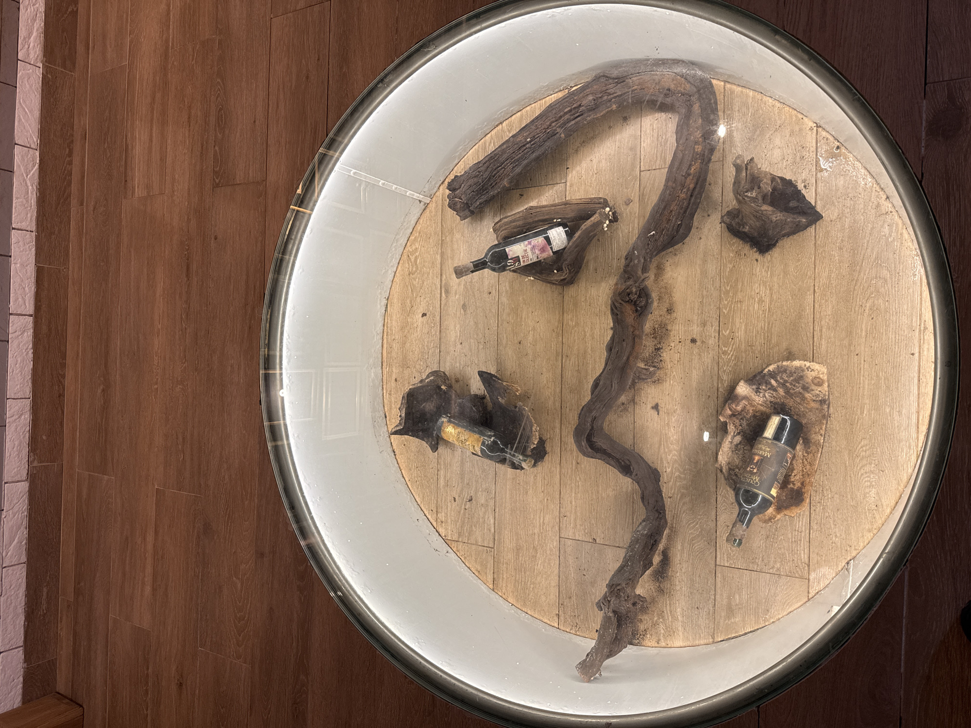 In a dimly lit room with wood-paneled walls, a circular glass-topped table displays a collection of dark, gnarled driftwood pieces arranged on a light wood floor. Three bottles are placed amongst the driftwood. One bottle, with a gold label, is partially obscured by a piece of wood. Another bottle, with a red and white label, stands upright on a piece of driftwood. A third bottle, with a dark label featuring the text "COLECIÓN MEDIO", lies on its side on a rough piece of driftwood. The table is illuminated by a ring of light around its perimeter. The floor outside the table is also wood-paneled. The scene is likely within a residential setting or a small business in Cojușna, Moldova.