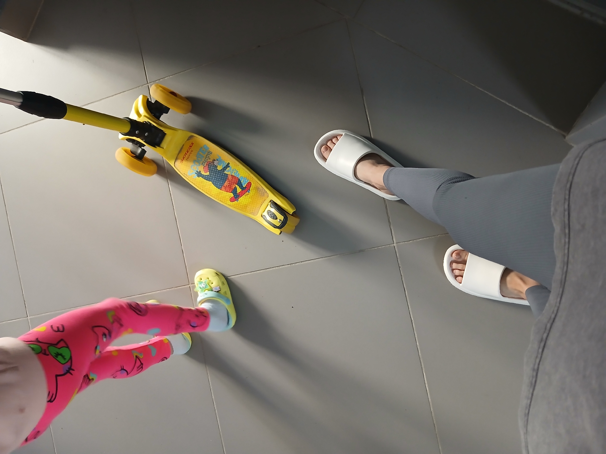 The image depicts a close-up, high-angle view of two pairs of feet on a tiled floor. One pair of feet, belonging to an adult, is wearing white slides and grey ribbed leggings. The other pair of feet, belonging to a child, is wearing bright pink leggings with cartoon designs and yellow clogs. To the left of the child's feet is a yellow three-wheeled scooter with cartoon graphics. The setting appears to be indoors, likely a home, given the tiled floor and the presence of children's toys and indoor footwear. The lighting suggests it could be daytime, as there are distinct shadows cast by the scooter and the feet. No specific weather or time of day is explicitly indicated, but the indoor setting implies the scene is not dependent on external conditions. The image composition focuses on the footwear and the scooter, suggesting an interaction or preparation for an activity involving these items, possibly a child about to ride the scooter. There is no discernible text in the image other than what appears to be on the scooter itself, which is too small to read clearly. The scene is domestic and casual, with no strong emotional cues conveyed.