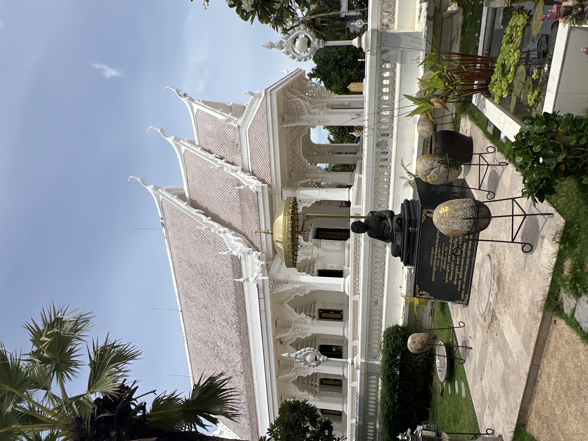 This is a medium shot of a white temple in Baan Bo Phut, Thailand, on a sunny day with a clear blue sky. In the foreground, there is a stone statue of a seated monk beneath a golden umbrella, surrounded by potted plants and large, egg-shaped stones on stands. A pond with lily pads is visible to the right. Behind the statue is a white ornate fence, and beyond that, the temple itself, with its distinctive multi-tiered white roof and intricately carved facade. Palm trees are visible to the left of the temple. The scene is serene and spiritual, likely a tourist attraction or place of worship.