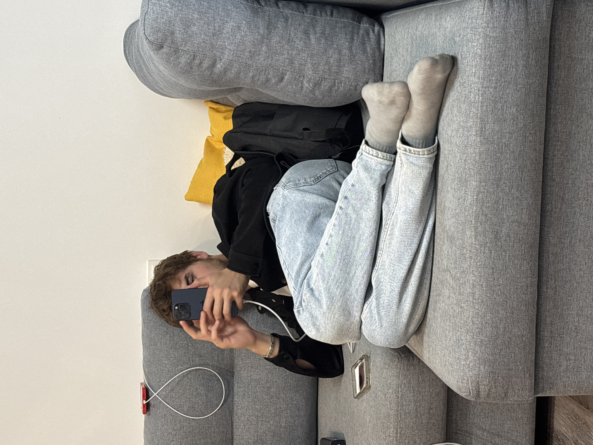 A young person with brown hair is lying on a gray sectional sofa, holding a dark blue smartphone in their hands and looking at it. They are wearing a black jacket over a white t-shirt and light blue jeans. The person's feet, covered in gray socks, are crossed and resting on the sofa. A black backpack is placed behind them on the sofa. Two yellow throw pillows are visible behind the person's head. A red charging brick with a white cable is on the backrest of the sofa to the person's left. A small clear case is on the seat of the sofa in front of the person. The environment appears to be indoors, with a plain white wall in the background.