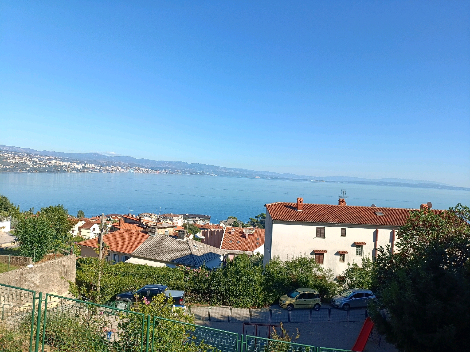 The image depicts a sun-drenched view of the city of Opatija, Croatia, overlooking the Adriatic Sea. In the foreground, a green chain-link fence runs along the bottom of the frame, with a red slide and playground equipment visible behind it. Several cars are parked on a paved area, including a black SUV, a yellow hatchback, and a silver sedan. Beyond the parking area, lush green trees and hedges separate the foreground from a cluster of residential buildings with characteristic red-tiled roofs. The buildings vary in size and style, with some featuring light-colored facades and dark-shuttered windows. The middle ground transitions into a panorama of the city of Opatija, characterized by its densely packed buildings extending along the coastline. The distant background is dominated by the calm, shimmering blue expanse of the Adriatic Sea, meeting a hazy, mountainous horizon under a clear, bright blue sky. The overall scene conveys a peaceful and picturesque coastal environment bathed in bright sunlight.