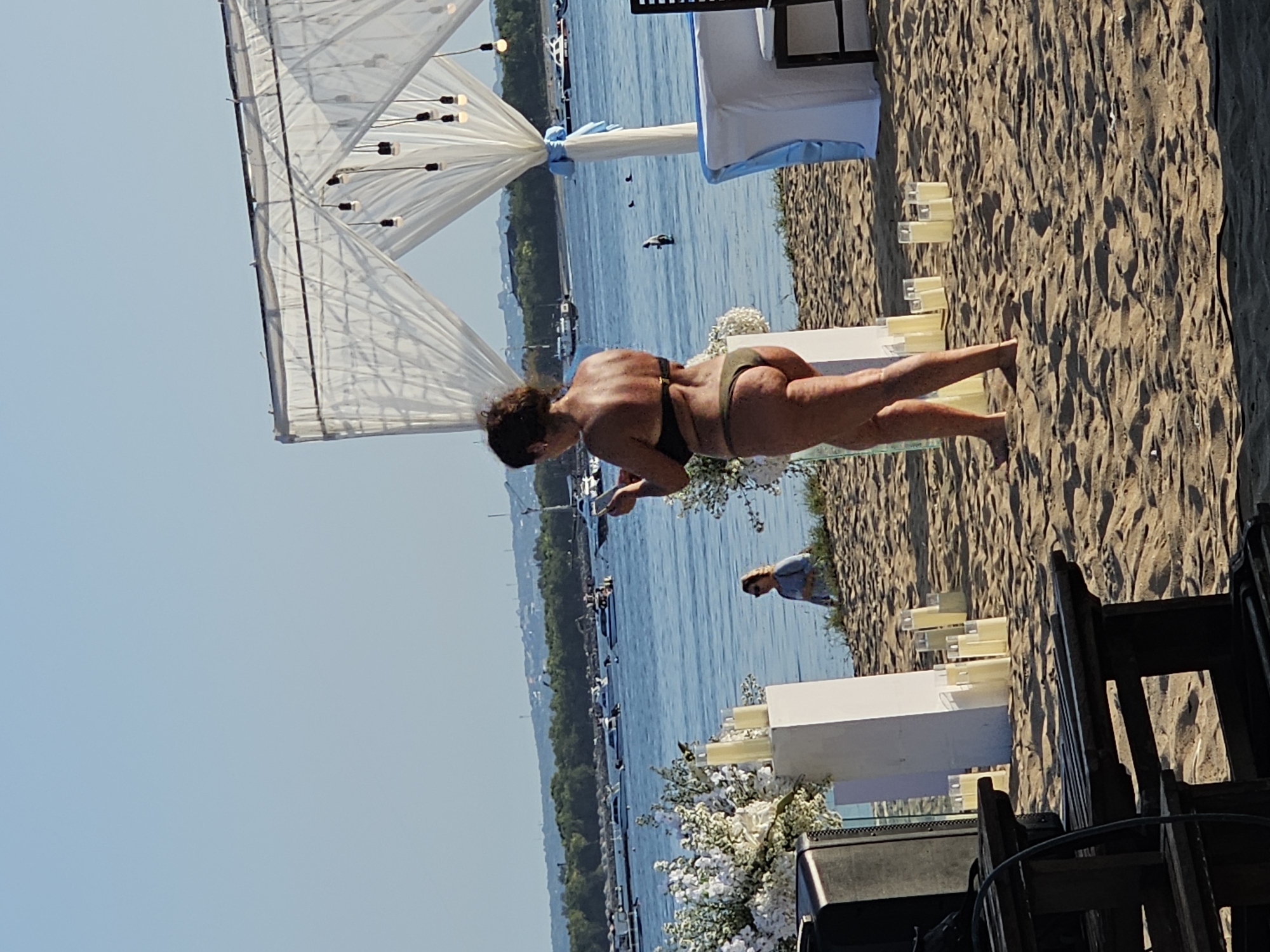 The image captures a beach scene. A woman is walking on the beach, wearing a dark-colored bikini. The background reveals a body of water with several boats and a shoreline with trees. A white tent-like structure stands on the beach, adorned with lights and flowers. There is an elaborate white floral arrangement, candles, and a fair-skinned person near the water. The location cannot be determined from the provided image and there is no way to confirm it is in Denpasar, Indonesia.